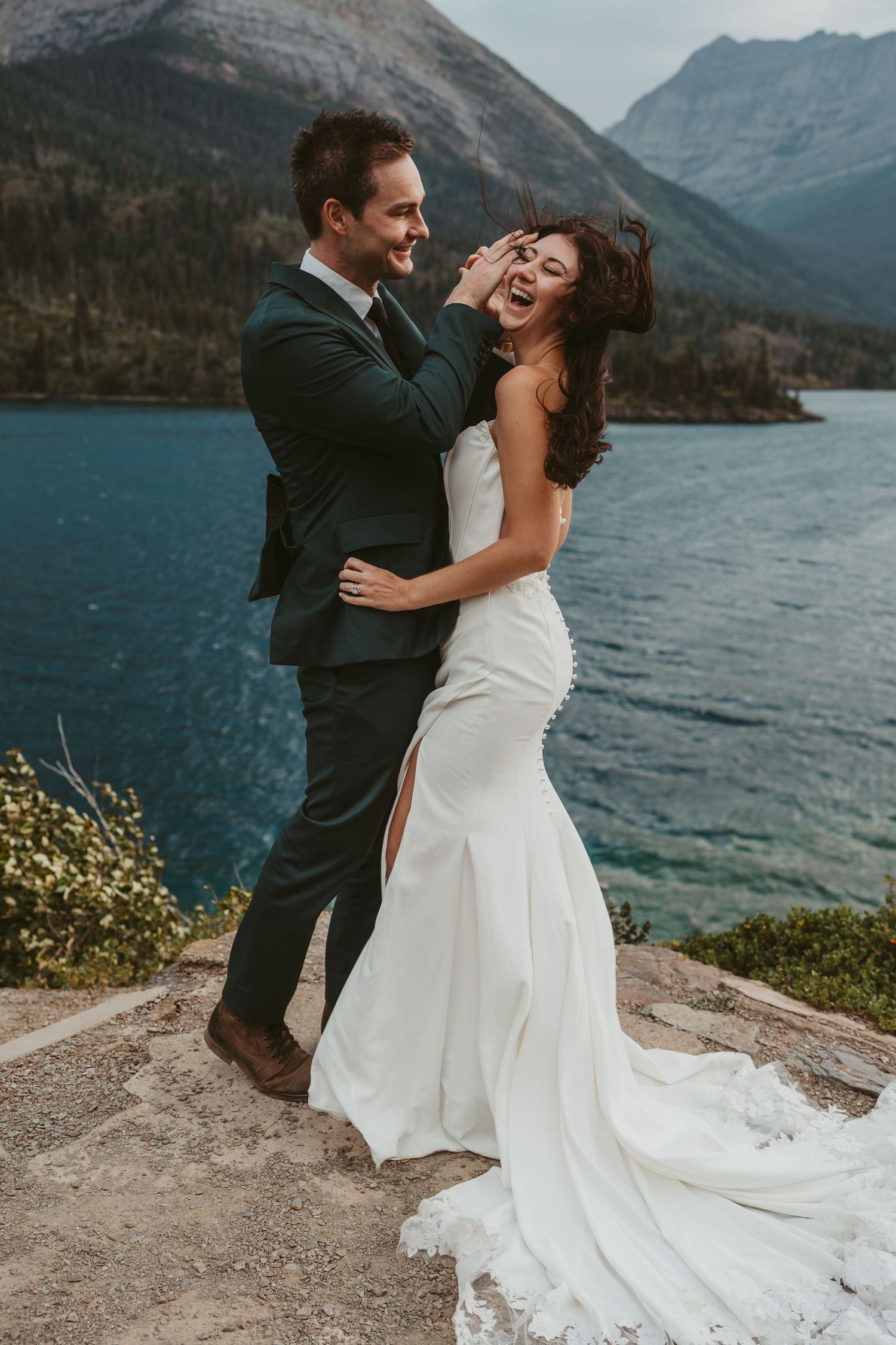 A joyful wedding couple embraces and laughs together on a rocky overlook with mountains and a lake in the background.