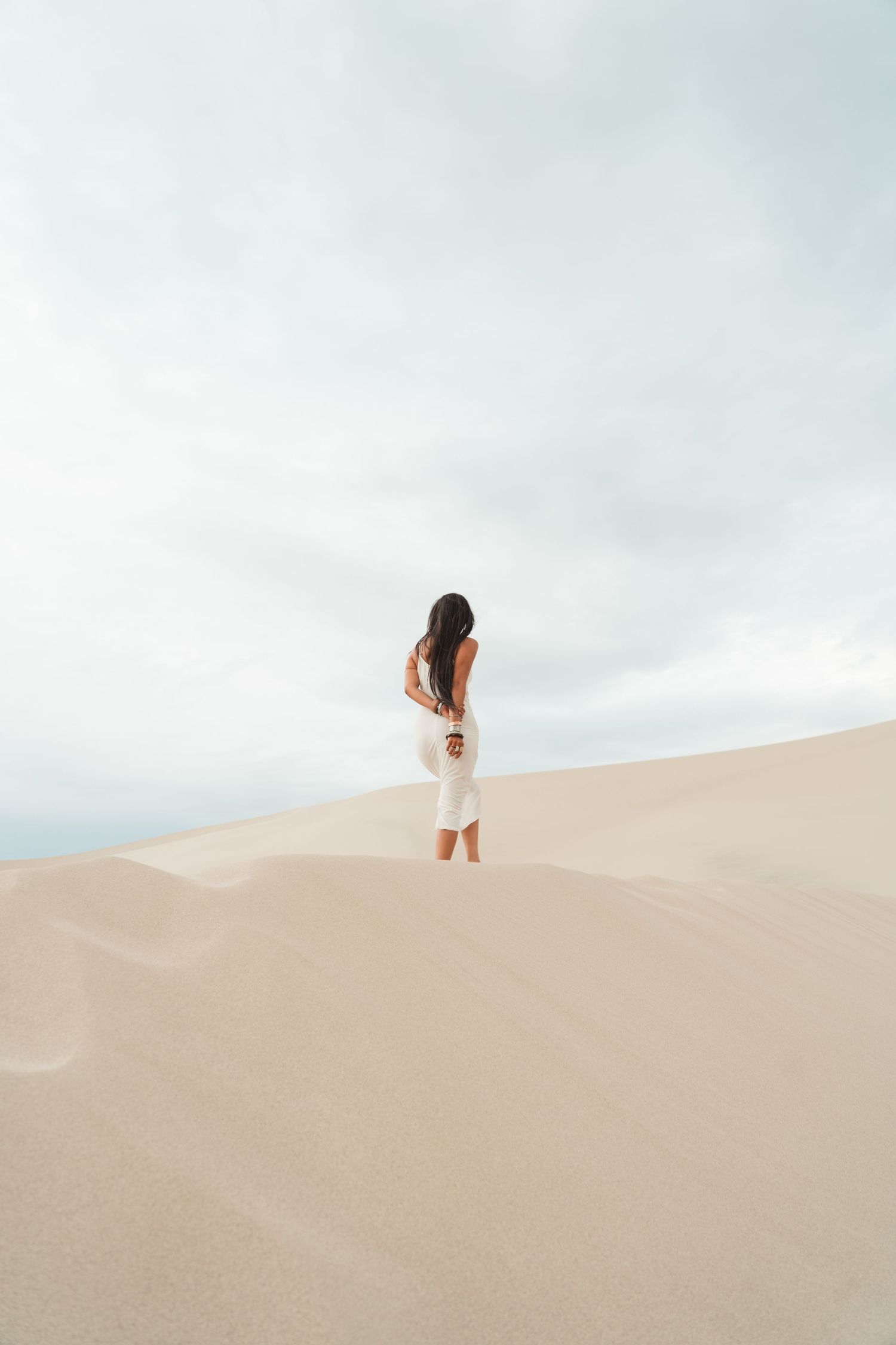 Woman in white dress standing on sandy desert dunes under cloudy sky.