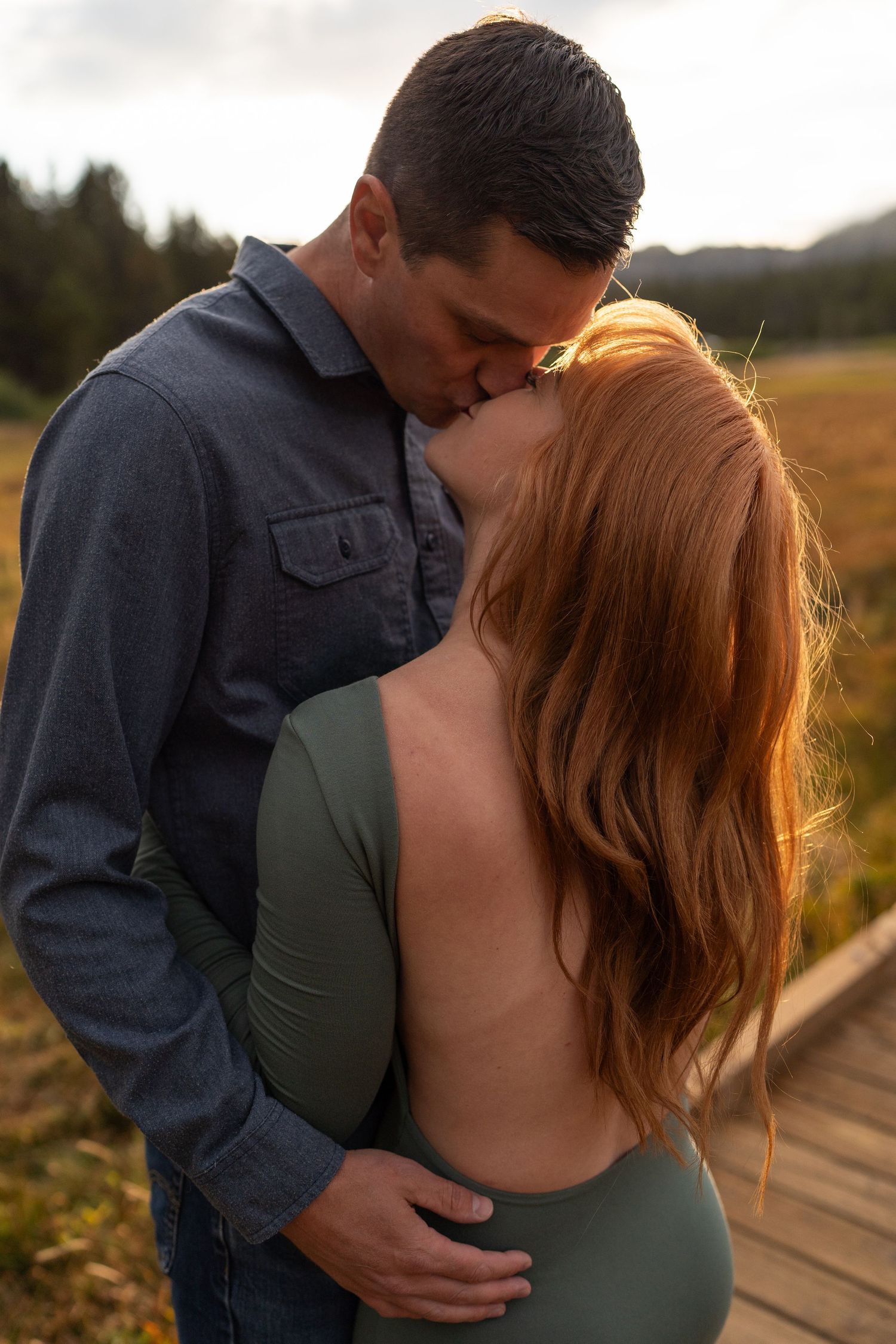 A couple shares a romantic kiss outdoors on a wooden path, the woman in a backless green dress with flowing red hair.