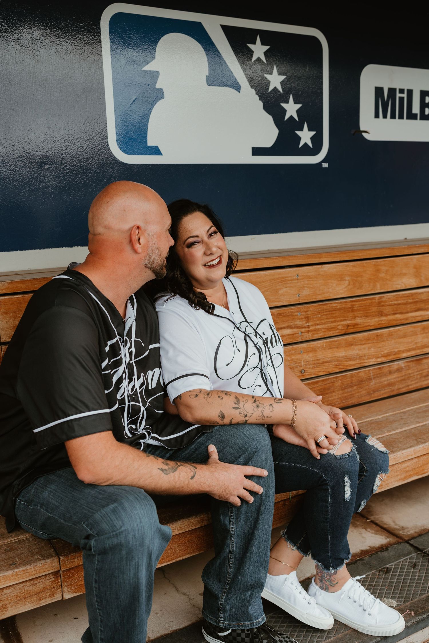 Couple smiling and sitting together in a baseball dugout, wearing jerseys, with an MLB logo on the wall behind them.