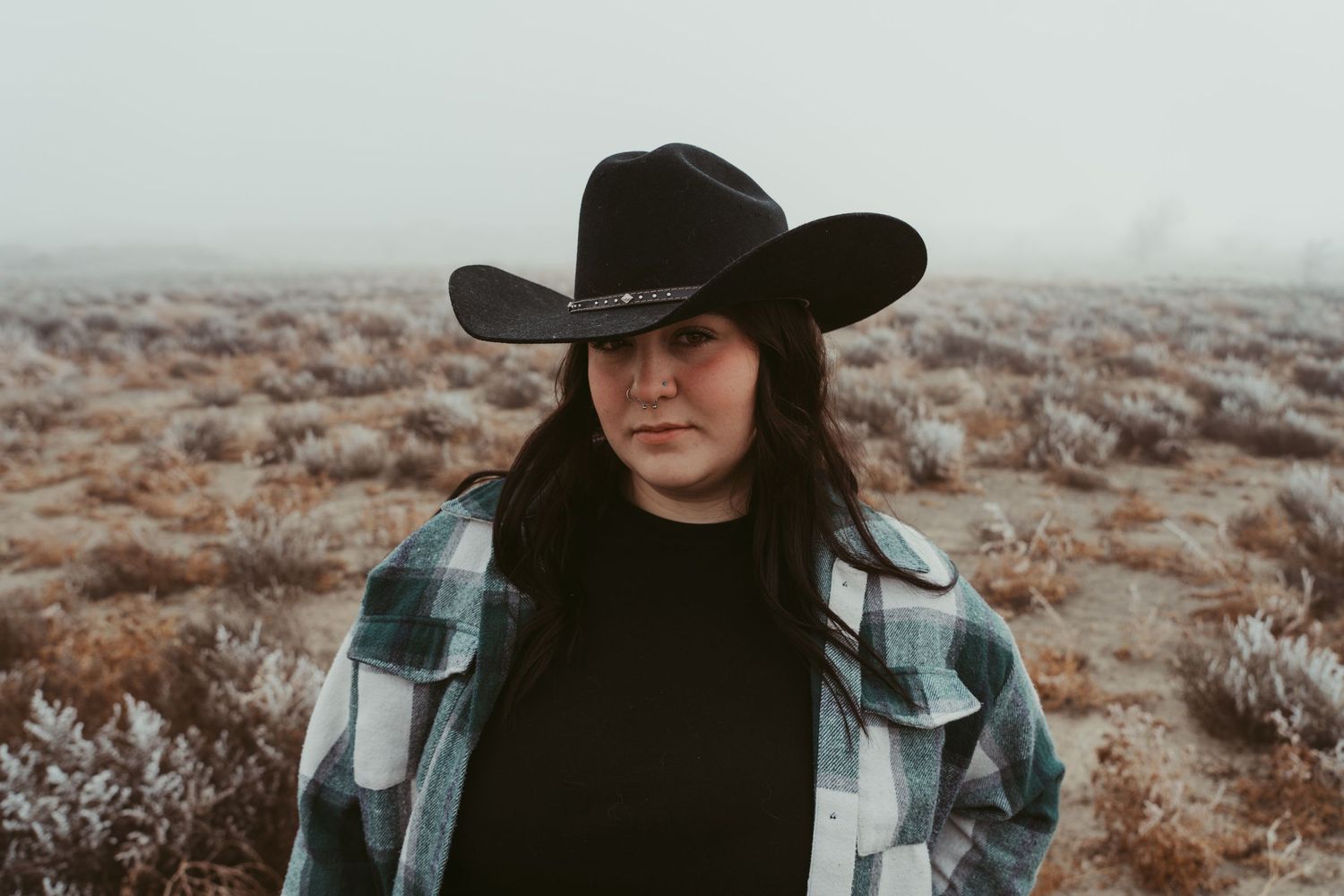 A cozy flannel shirt and black cowboy hat are worn against a foggy desert landscape in moody lighting.