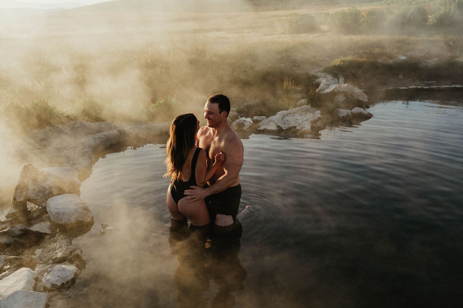 A couple embraces in steamy hot springs surrounded by rocky formations at sunset.