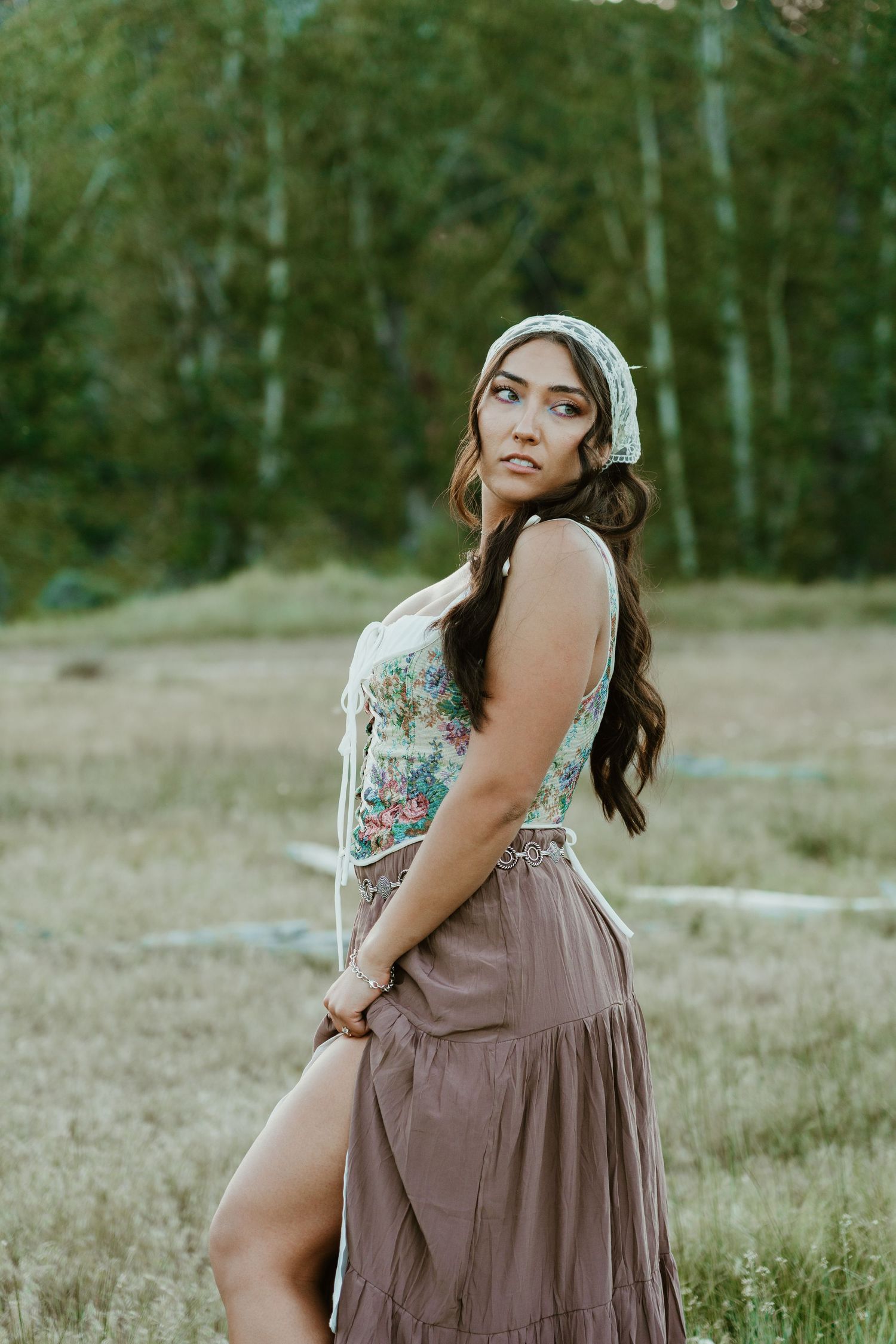 Young woman in floral top, brown maxi skirt, and white headscarf poses gracefully in a sunlit meadow with trees behind her.
