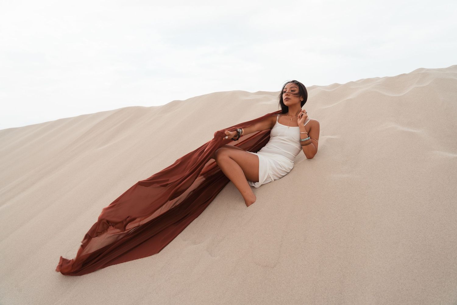 Woman in white dress with flowing brown fabric poses gracefully on sandy desert dunes under overcast sky.