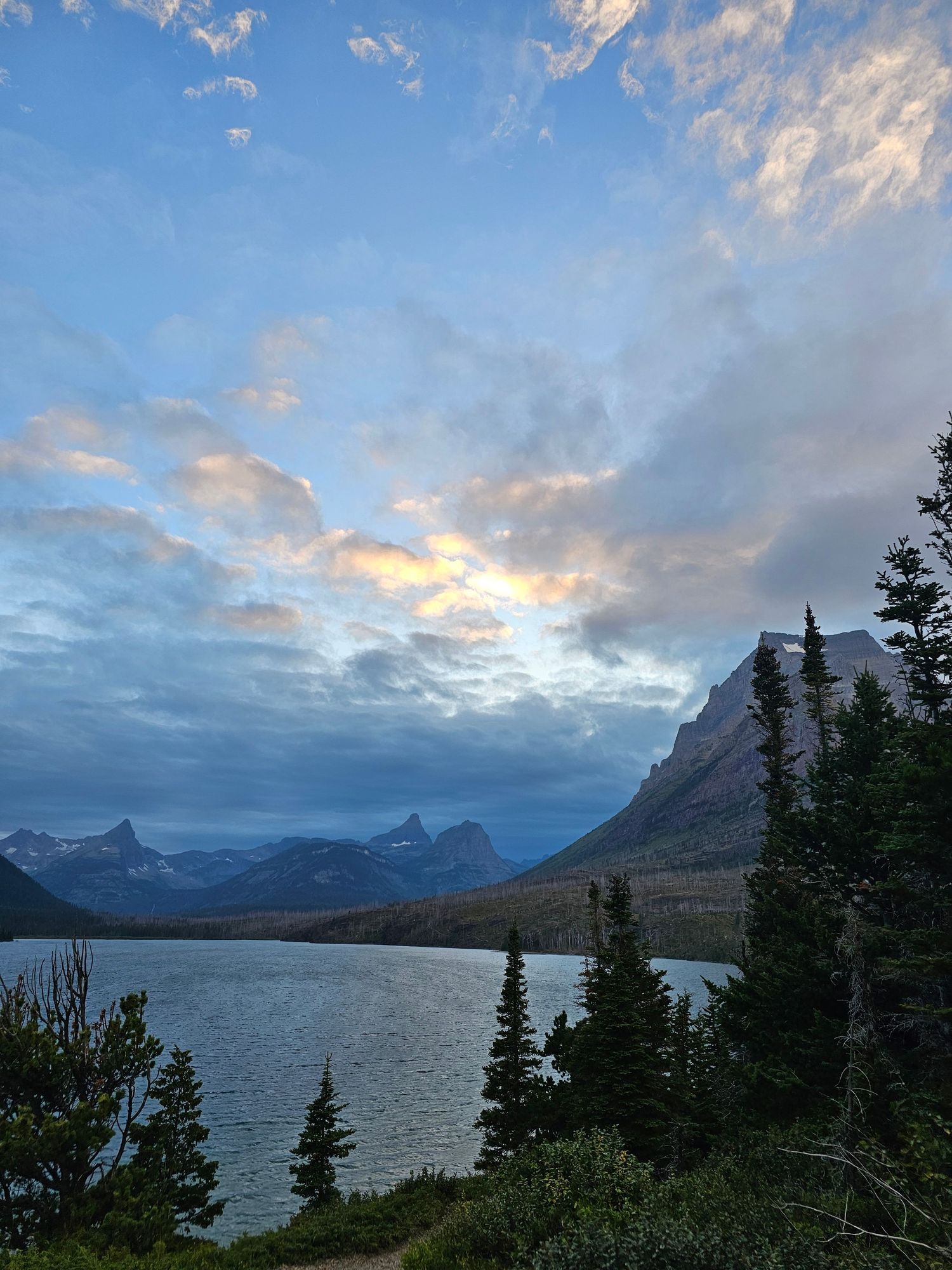 Scenic mountain lake vista in Glacier National Park with dramatic clouds and evergreen trees at sunset.