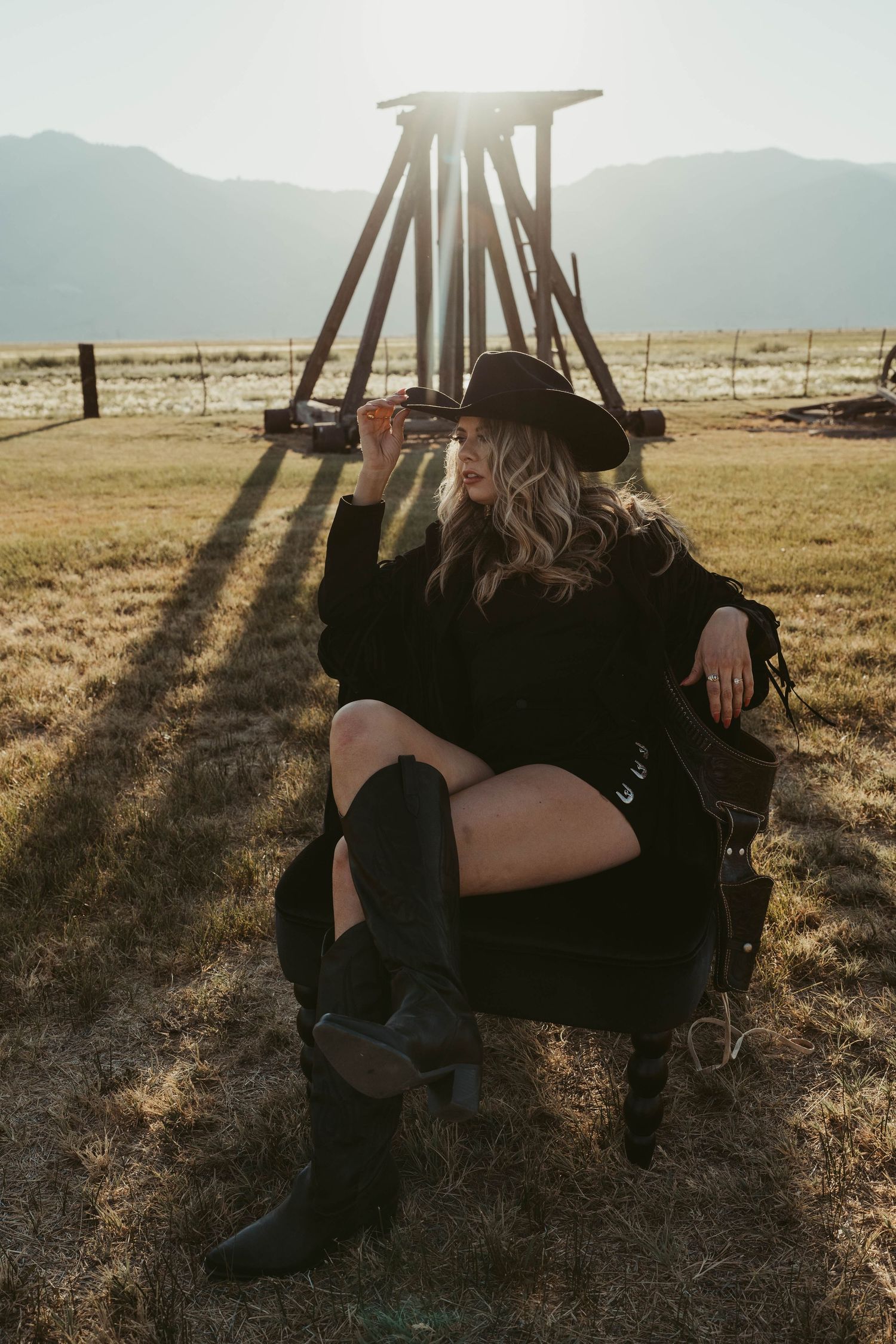 Silhouetted figure in western attire sits in field at sunset with tall wooden structure casting dramatic shadows.