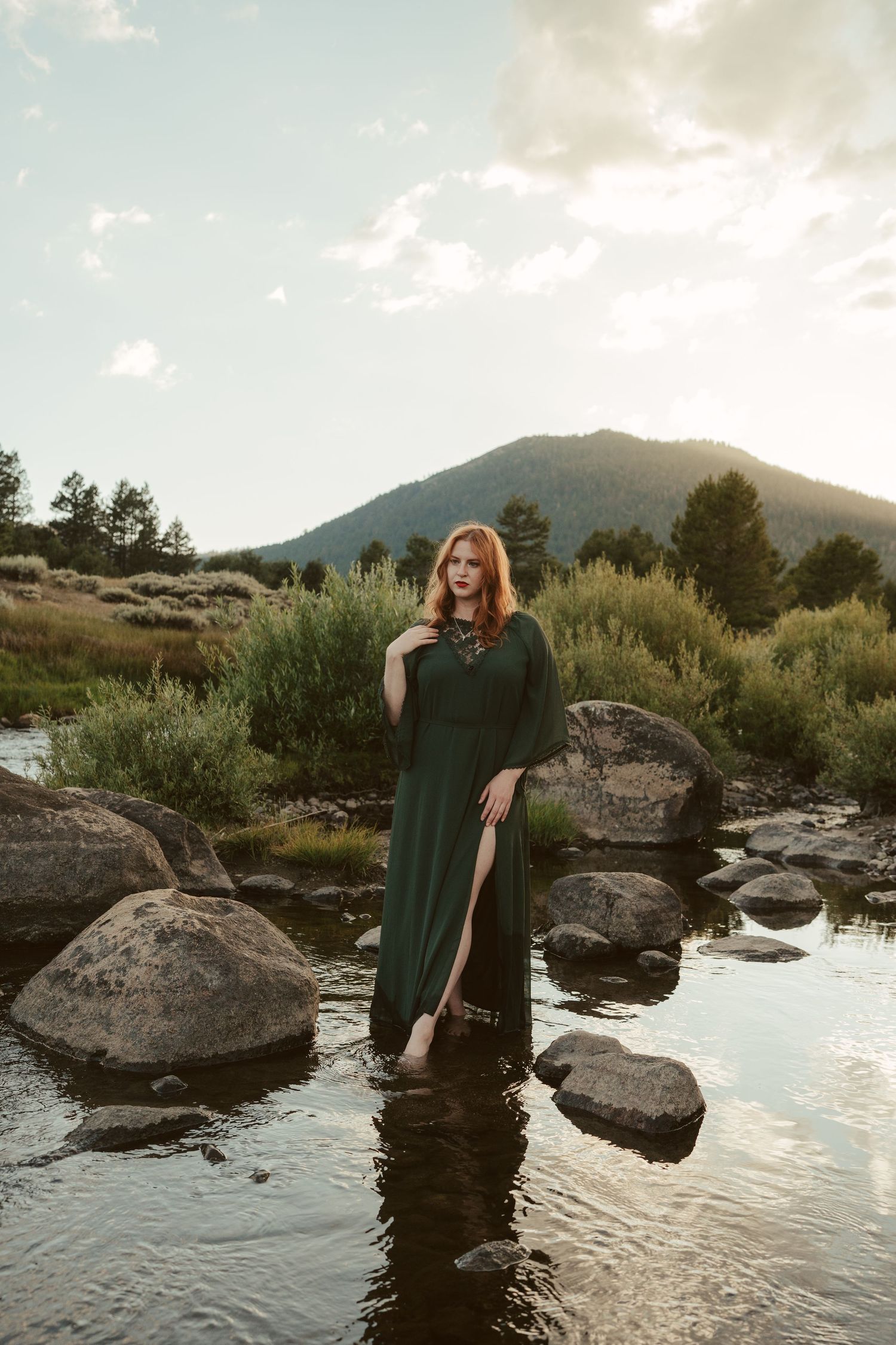 A person in a dark dress poses on rocks near water at sunset with mountains in the background.