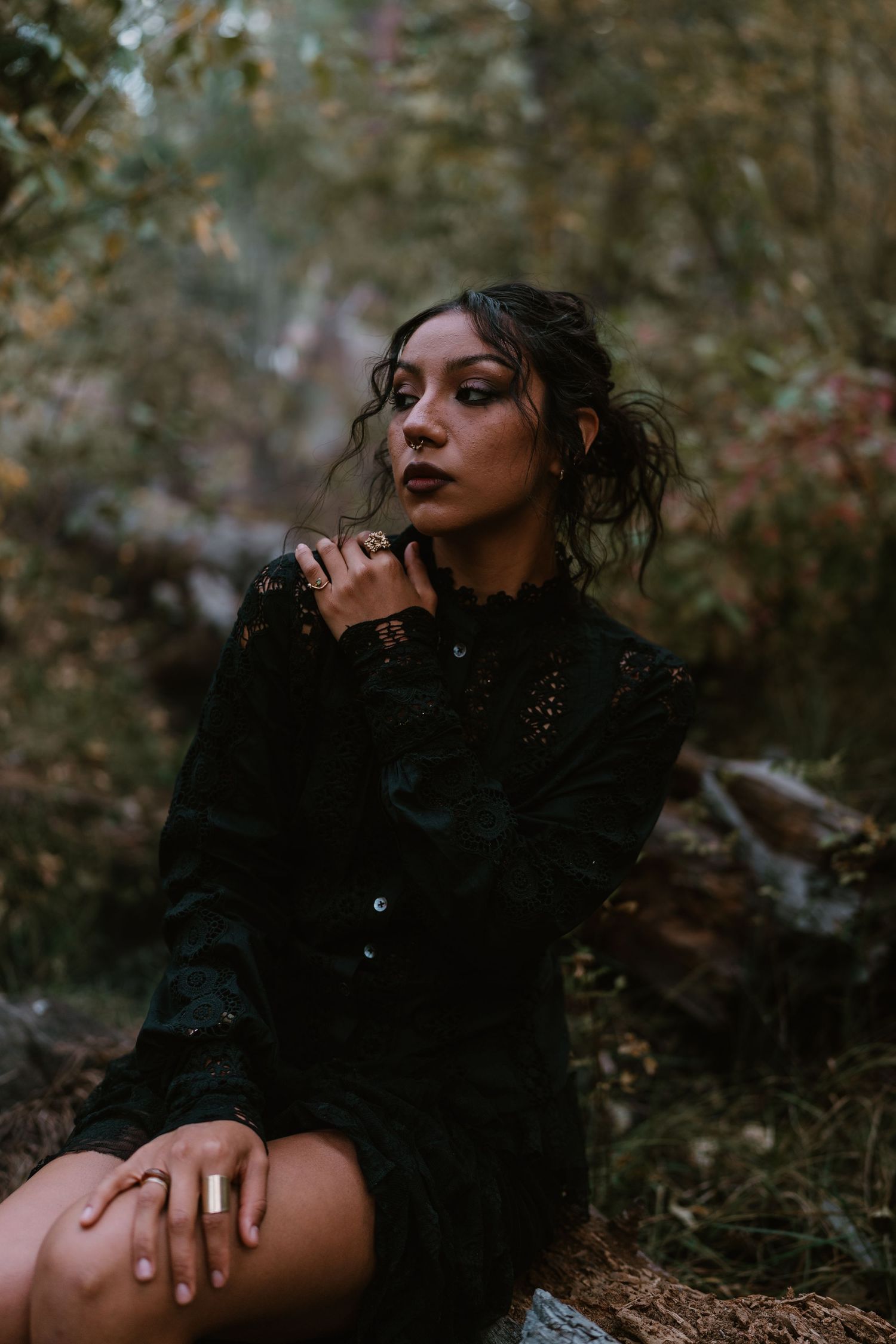 A woman in a black lace dress sits outdoors in a forest, wearing dark makeup and rings, gazing thoughtfully to the side.