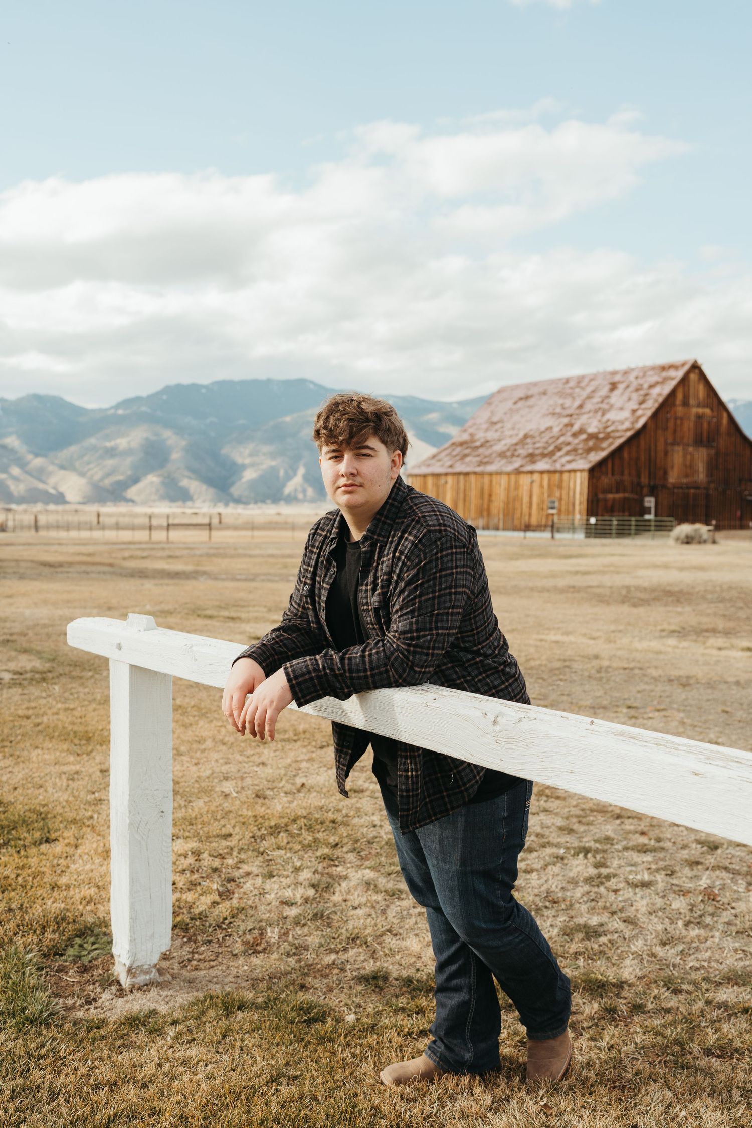 Person in dark sweater leans against white fence with rustic barn and mountains in background.