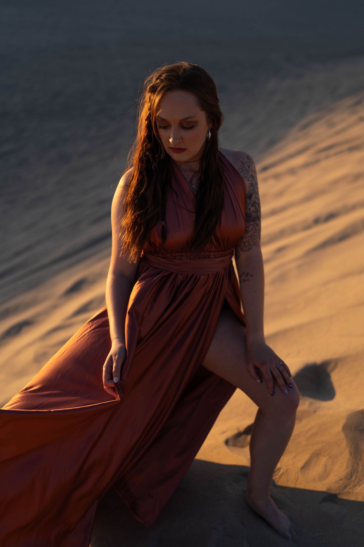 A flowing rust-colored dress catches the golden light against sandy dunes at sunset.