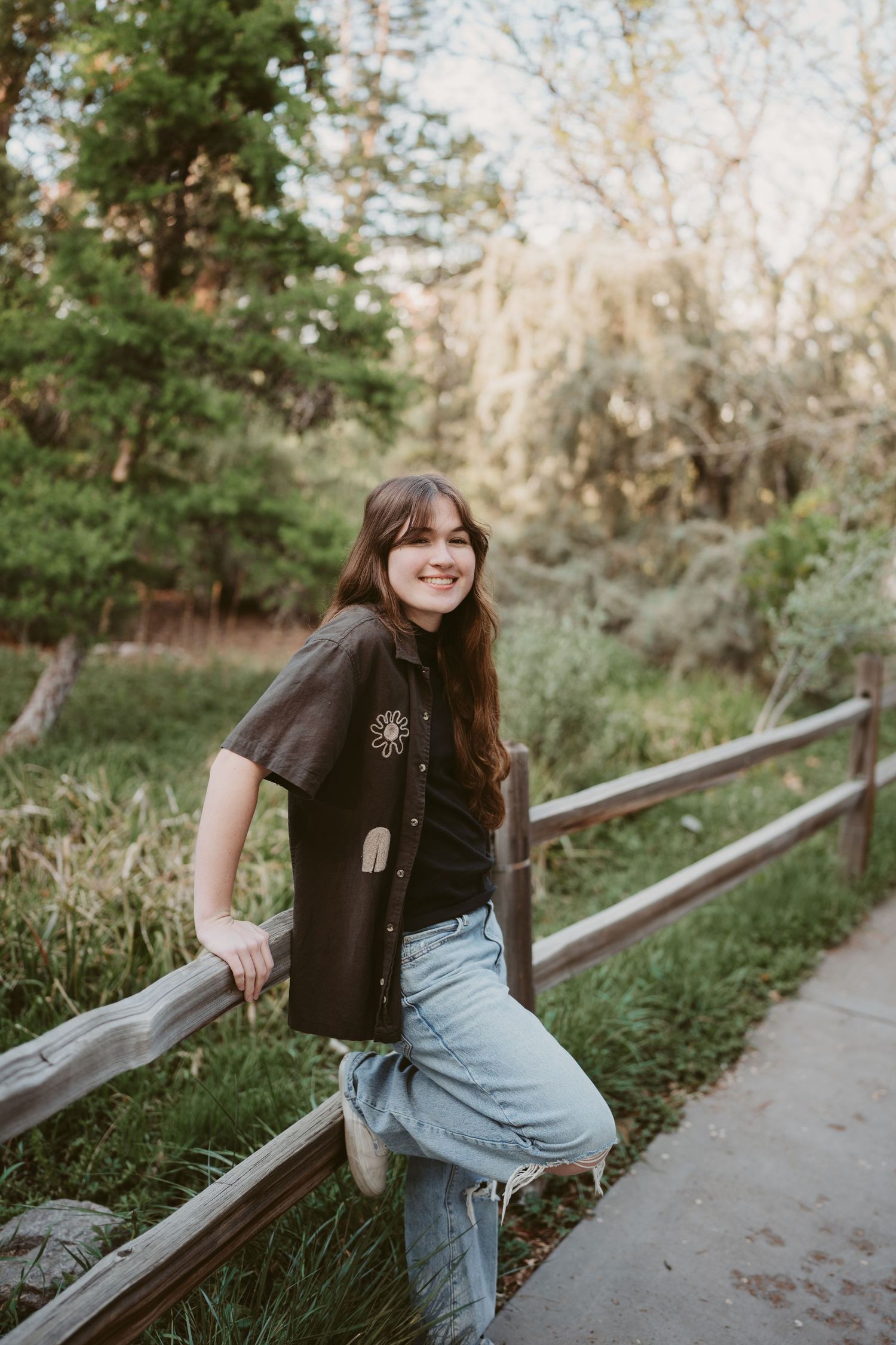 A person in a black top and jeans sits casually on a wooden railing along a tree-lined path on a sunny afternoon.