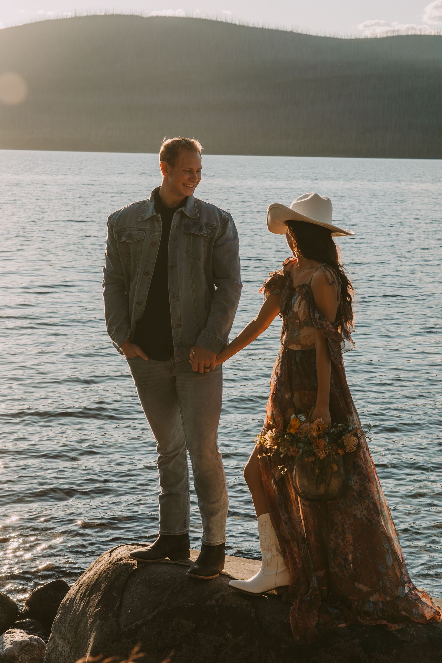 A couple stands on rocky shoreline at sunset enjoying scenic views of a serene lake in the background.