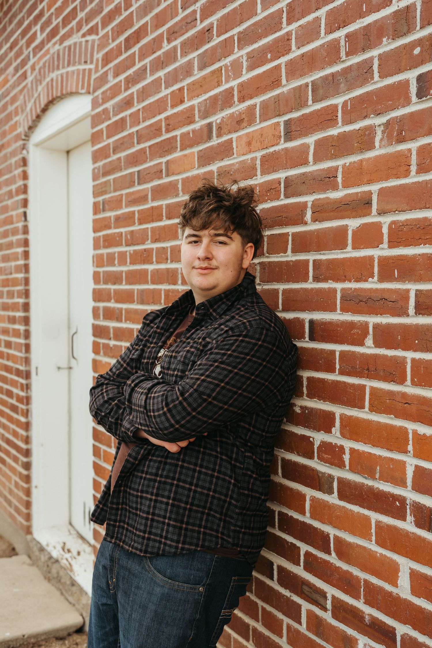A person in a plaid shirt and jeans leans against a brick wall with white-framed windows.