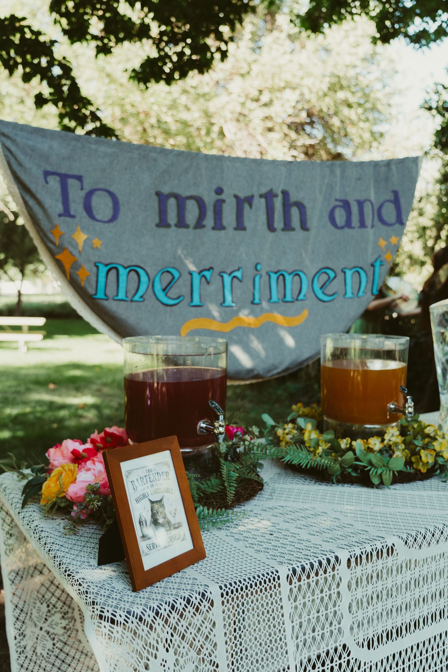 Outdoor party table with punch dispensers and a banner reading To Mirth and Merriment in a sunny garden setting.