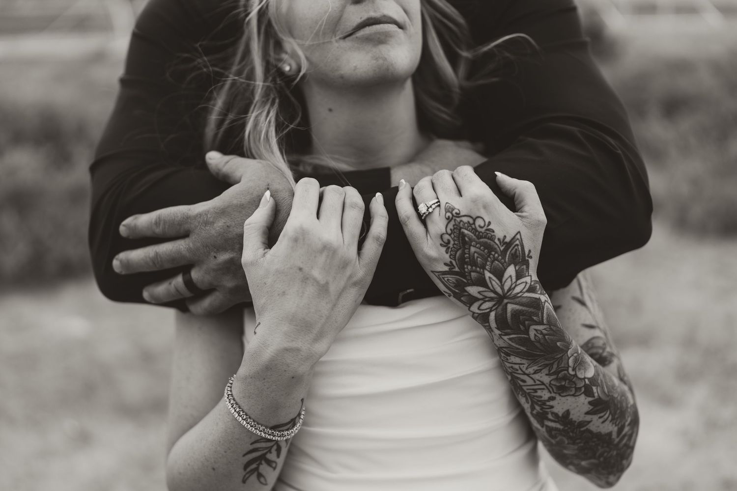 Black and white photo of a couple embracing, woman with floral sleeve tattoos and bracelet, man holding her from behind.
