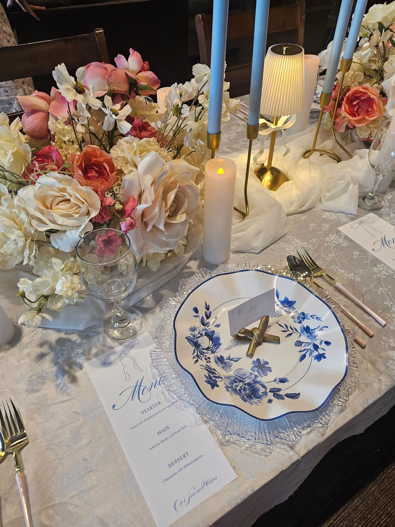 Elegant table setting with blue and white china plates, pink and white floral arrangements, and white lace tablecloth.