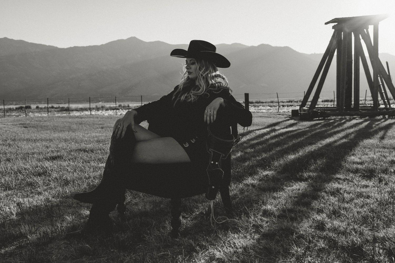 Black and white photo of someone in western attire sitting in an armchair outdoors with mountains in the background.