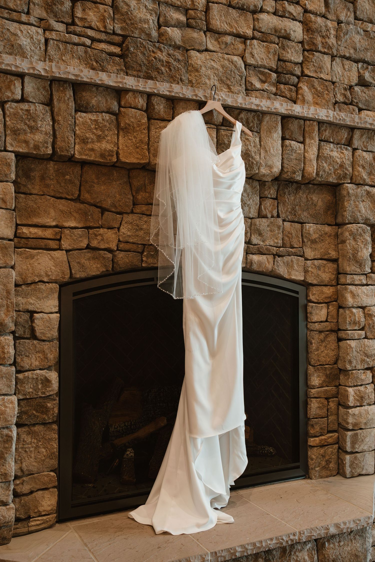 Elegant white satin wedding dress with veil hanging on a wooden hanger over a rustic stone fireplace.