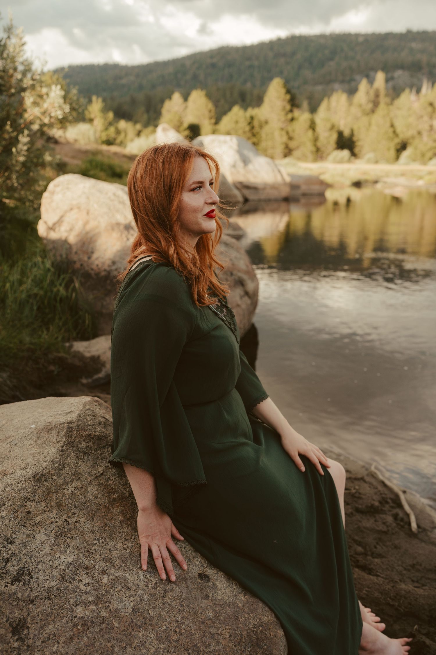 A woman in a dark green dress poses on rocks by a river with golden trees reflecting in the water.
