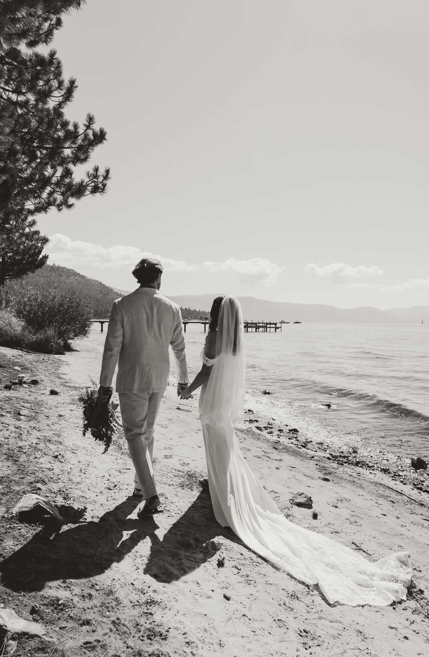 A couple in white attire walks along a scenic beach shoreline with mountains visible in the distance.