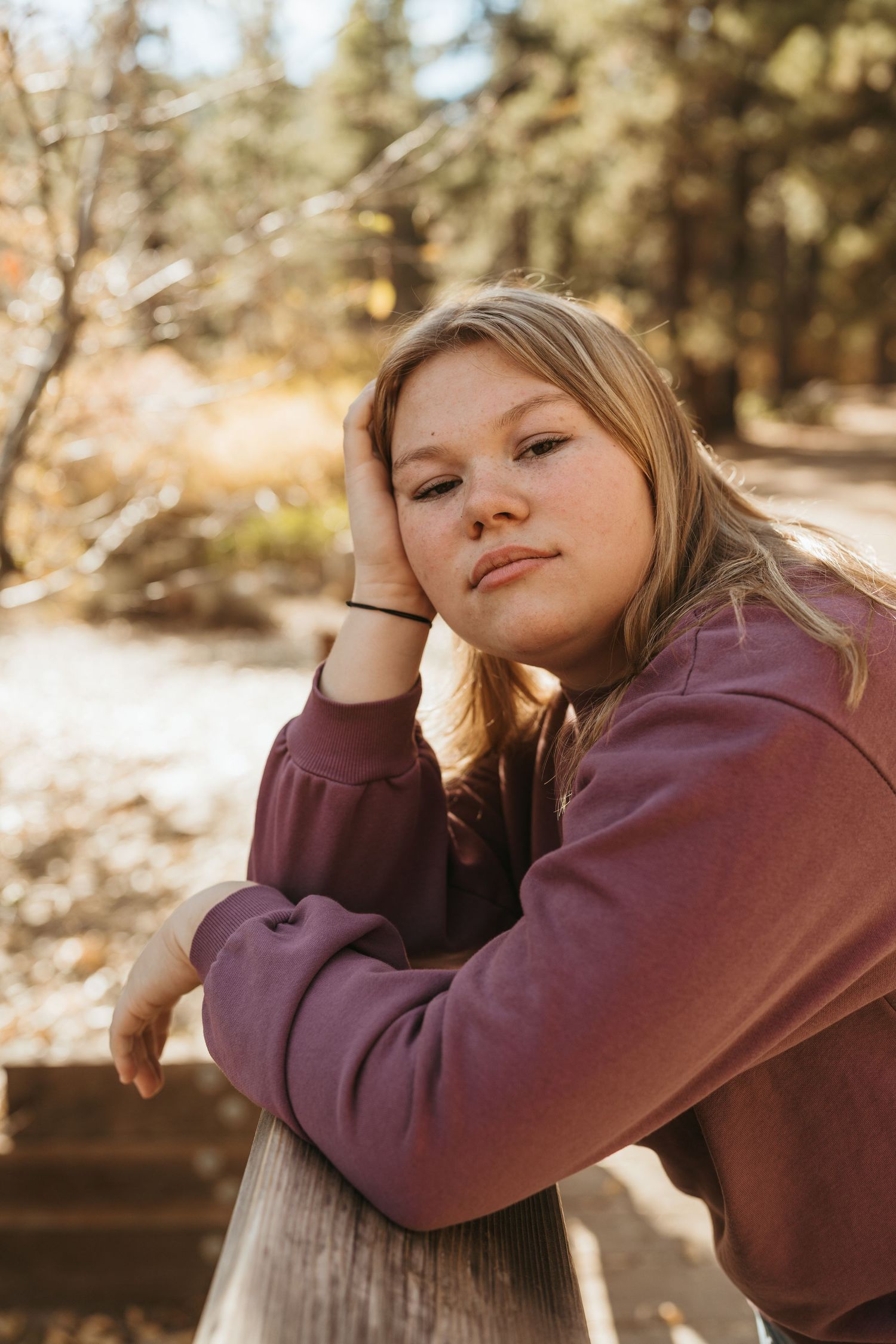 A person in a burgundy sweatshirt rests their head thoughtfully on a wooden railing in an outdoor setting.