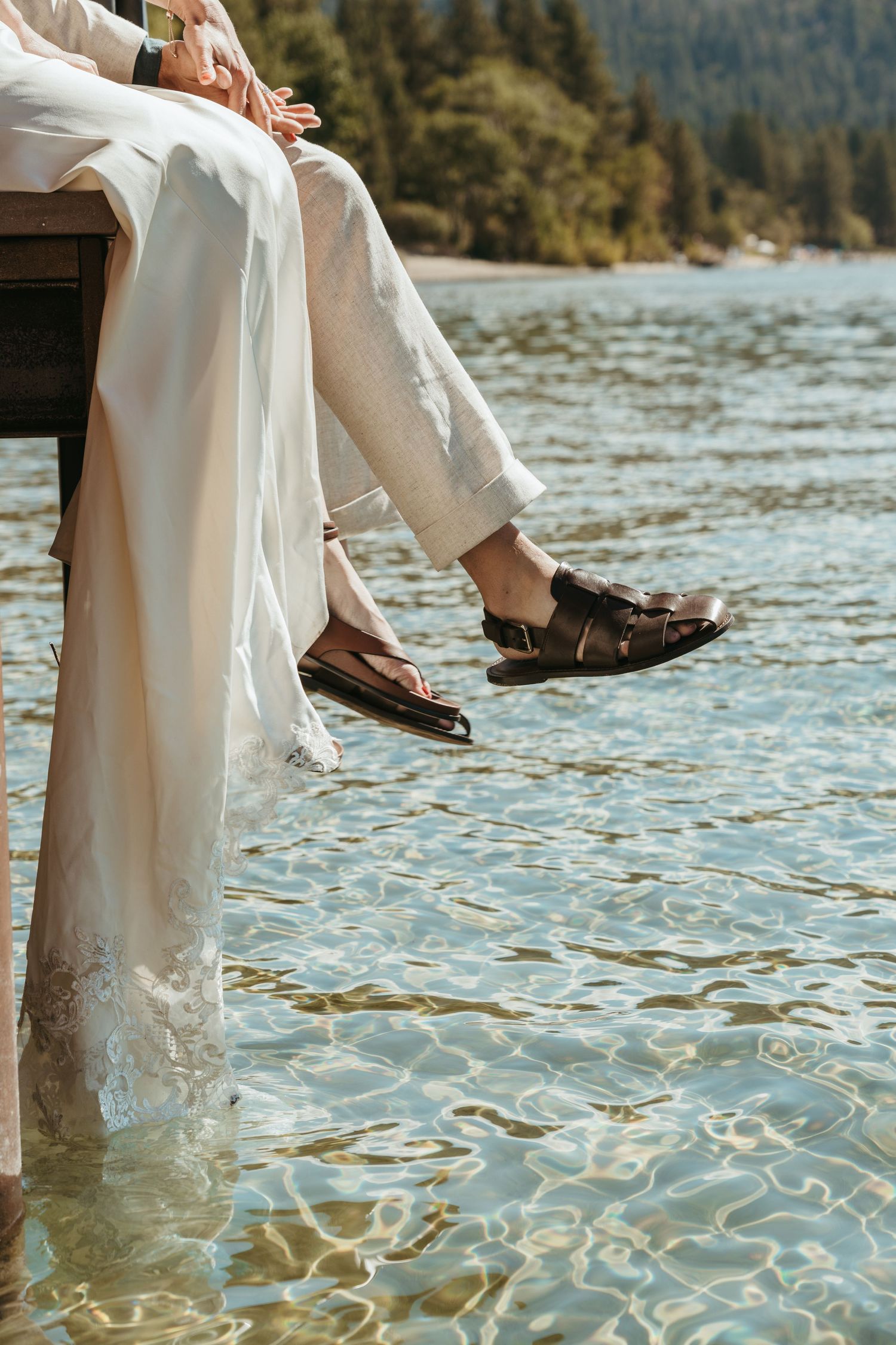 Two people in white clothing dangle their feet over a dock into sparkling lake water on a sunny day.
