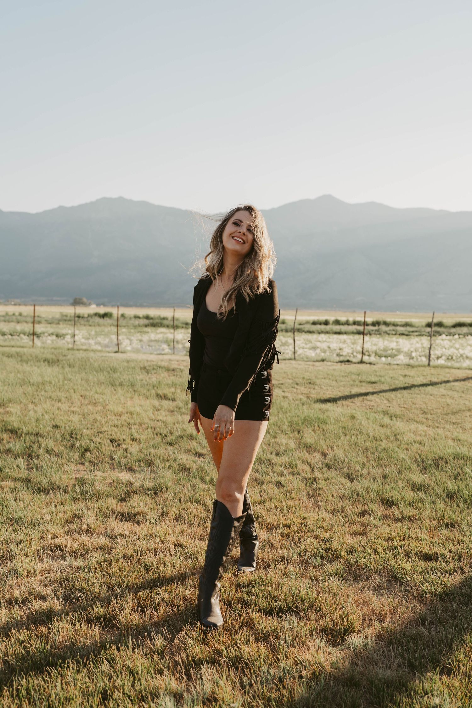 Person in black fringe jacket and tall boots stands in sunlit meadow with mountains in background on sunny day.