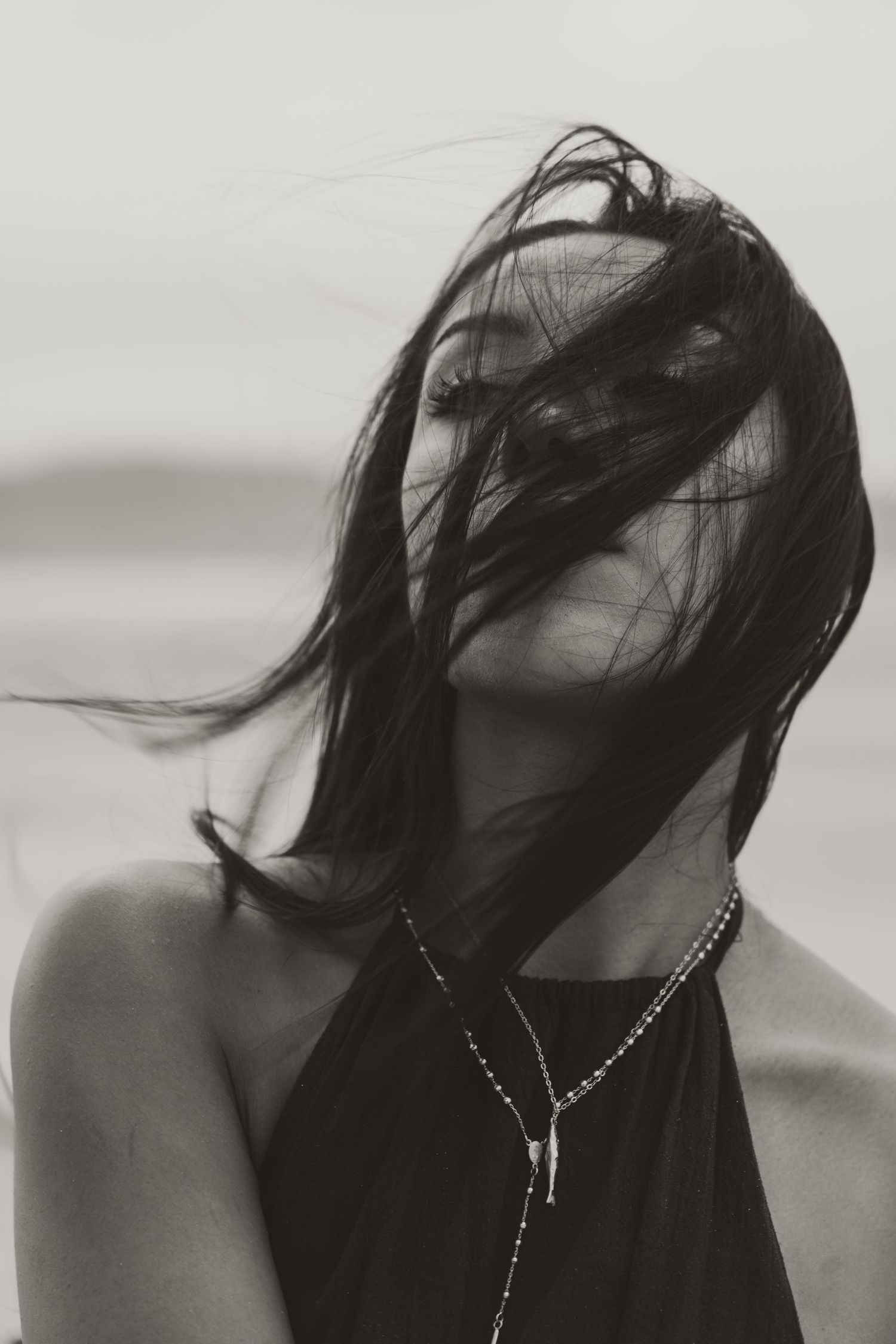 Black and white photo of a woman with dark hair blowing across her face in the wind, wearing a delicate necklace.