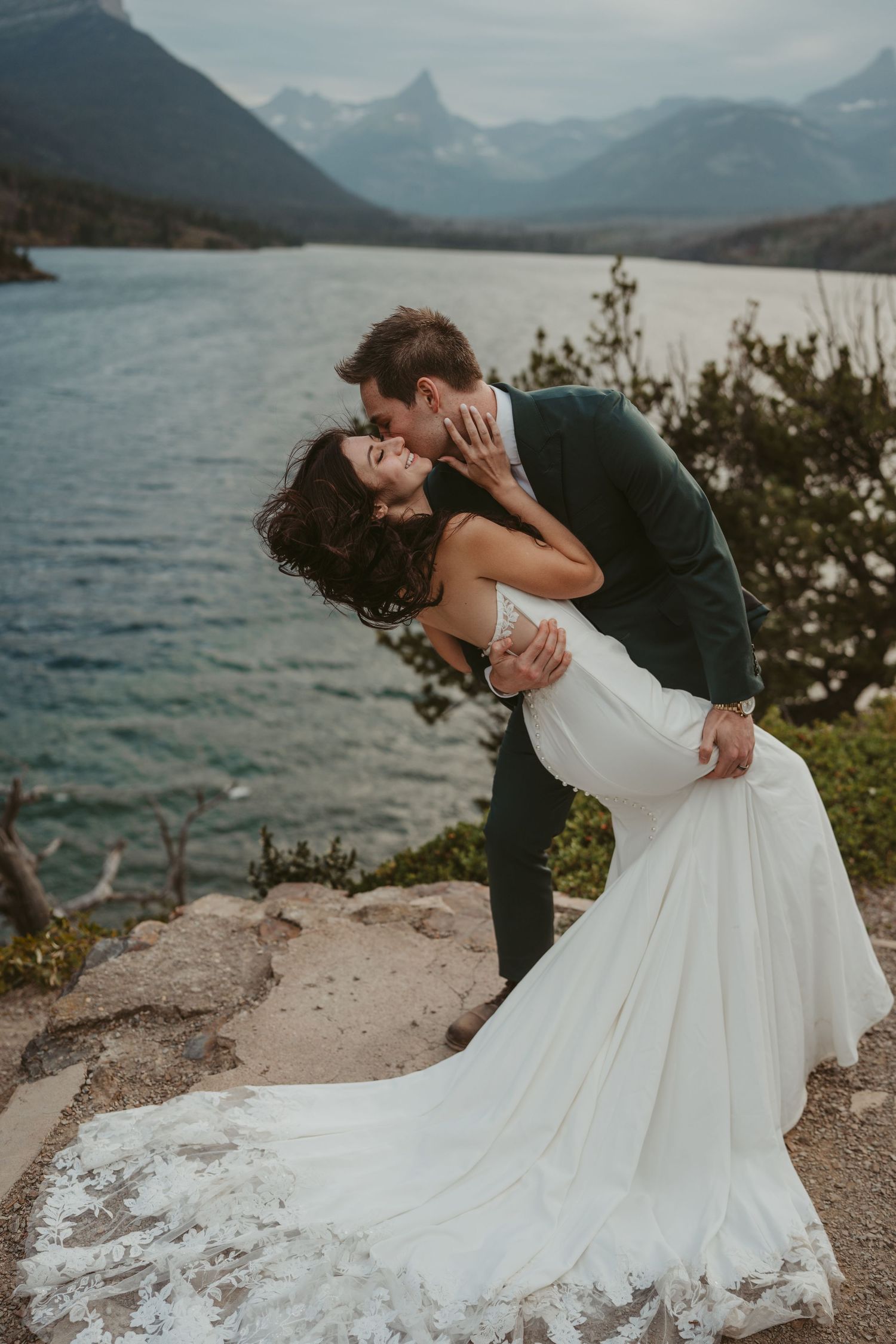 A romantic couple in wedding attire share an intimate moment on a rocky overlook with mountains and a lake in the background.