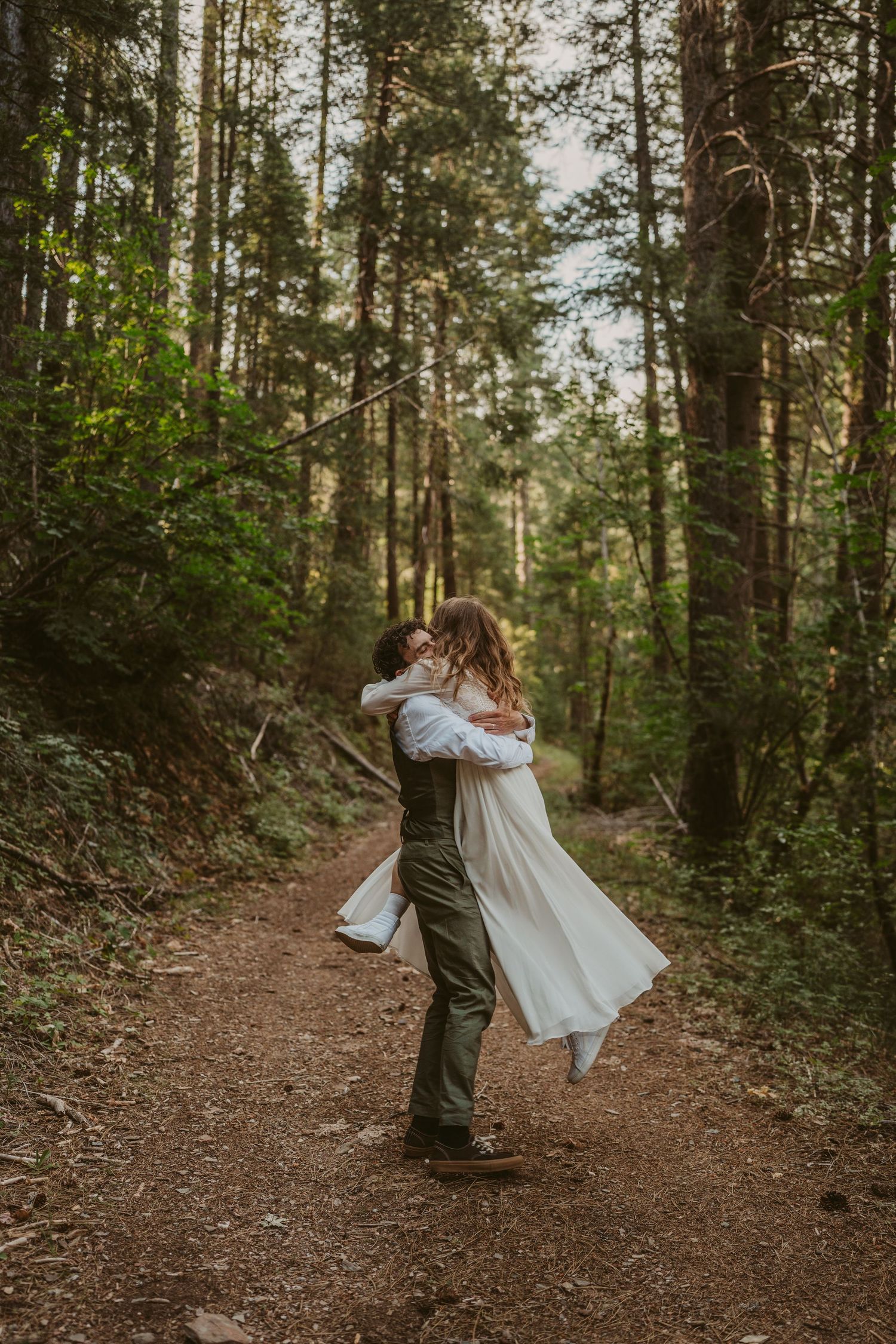 A couple embrace on a forest trail as sunlight filters through tall pine trees in the background.
