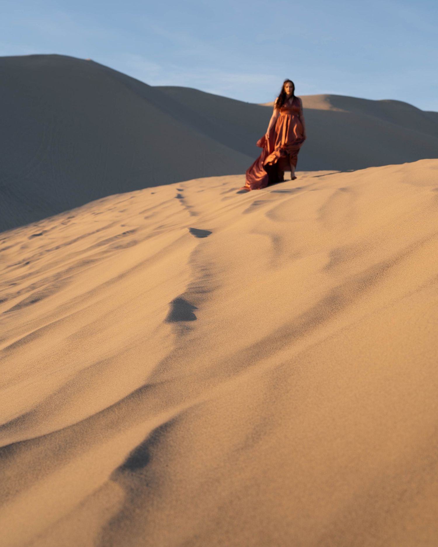 Figure in flowing red attire stands on golden sand dunes against a mountain backdrop during sunset.