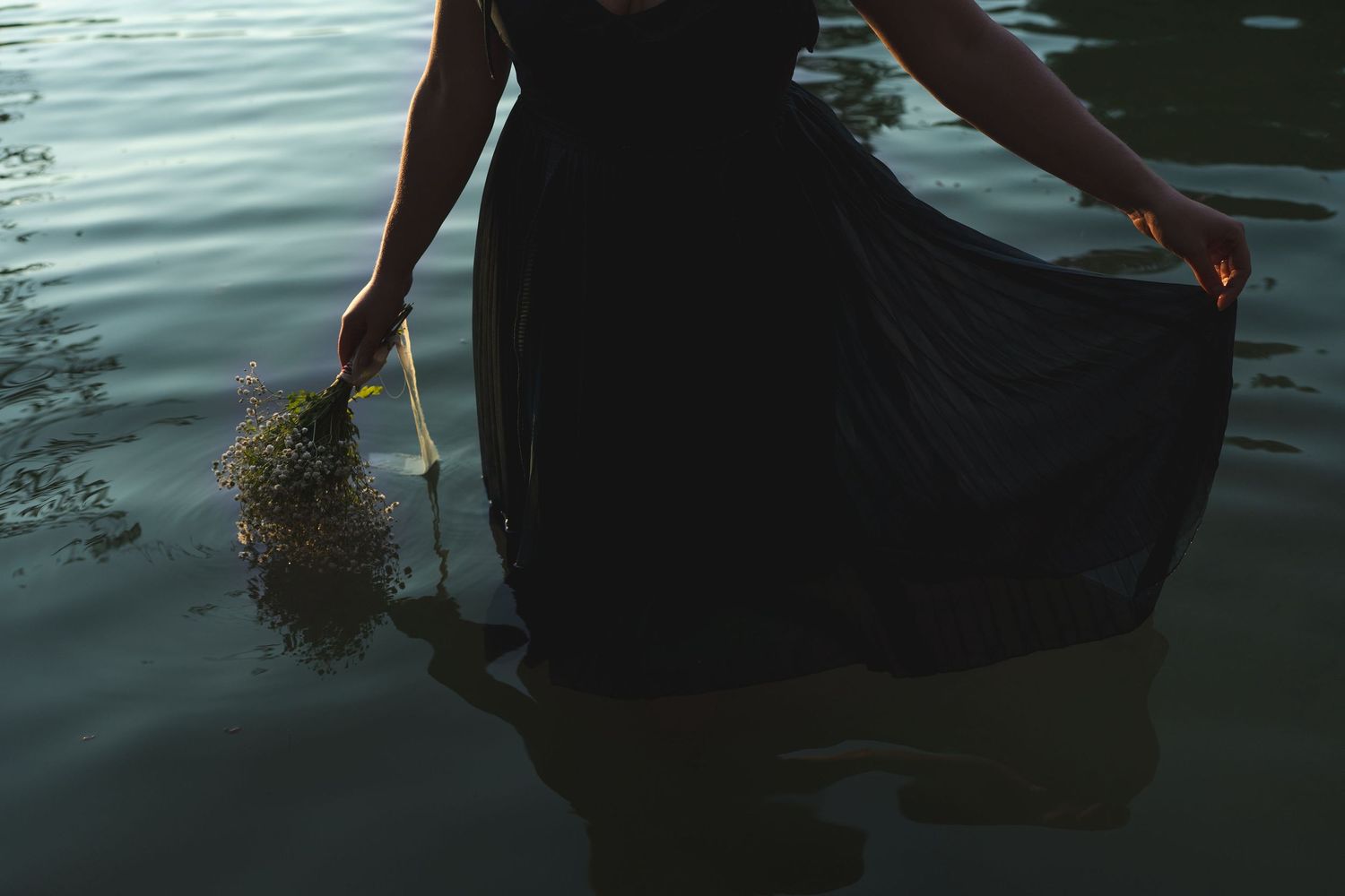 Woman in black dress standing in water holding a bouquet of flowers, moody atmospheric photography.