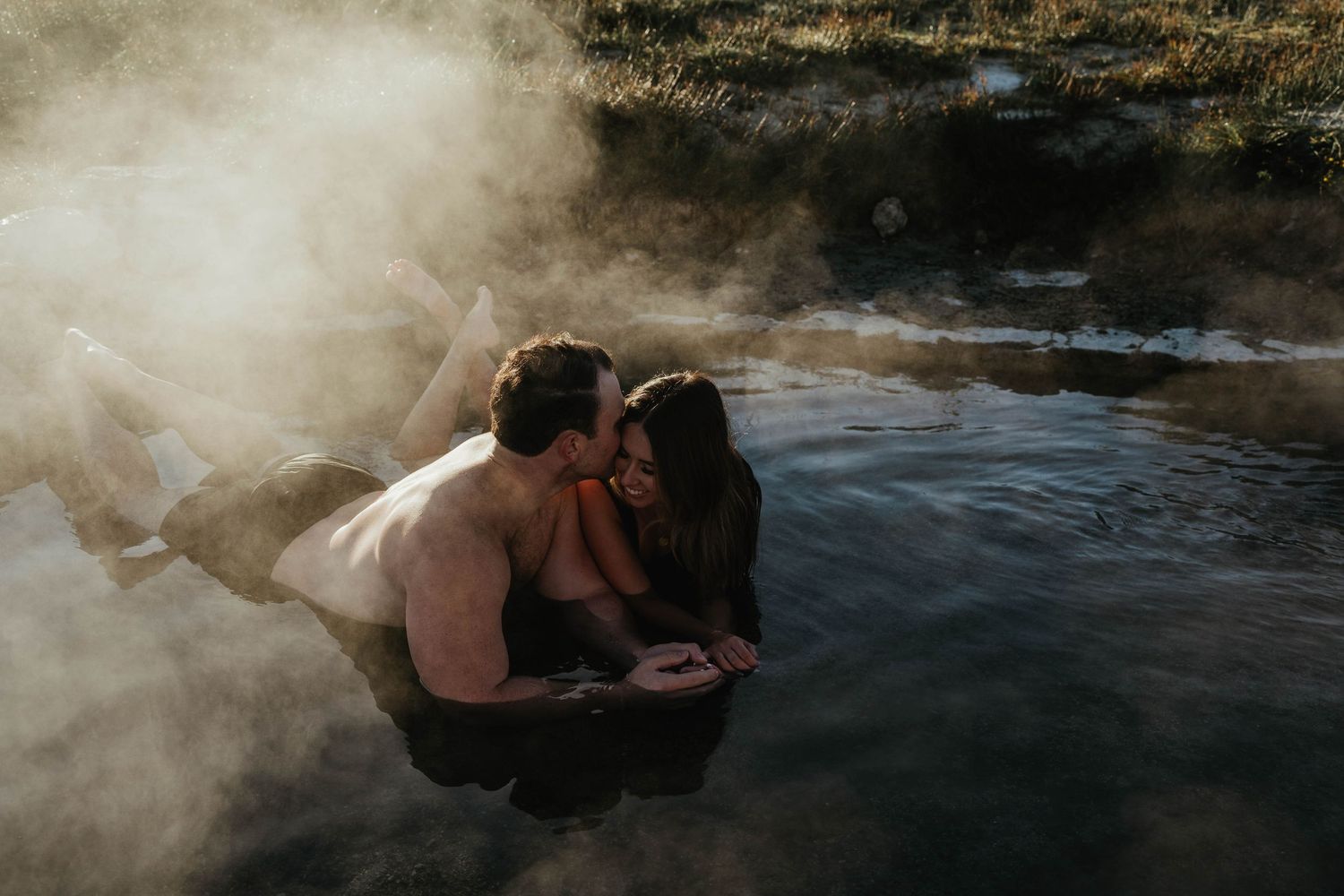A couple relaxes in a steamy natural hot spring surrounded by mist and rocks in a romantic outdoor setting.