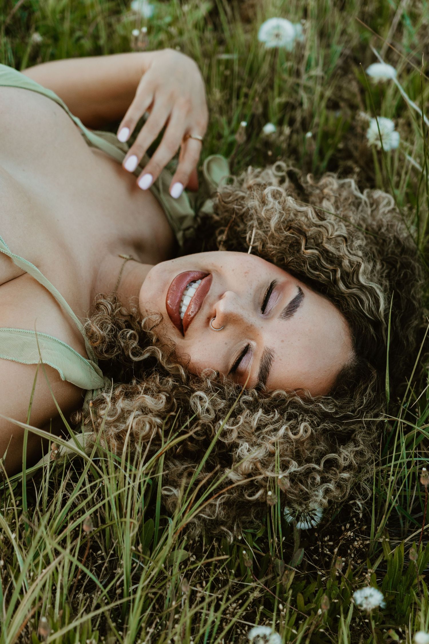 A smiling woman with curly hair lying in a green field of dandelions, wearing a sage green top.