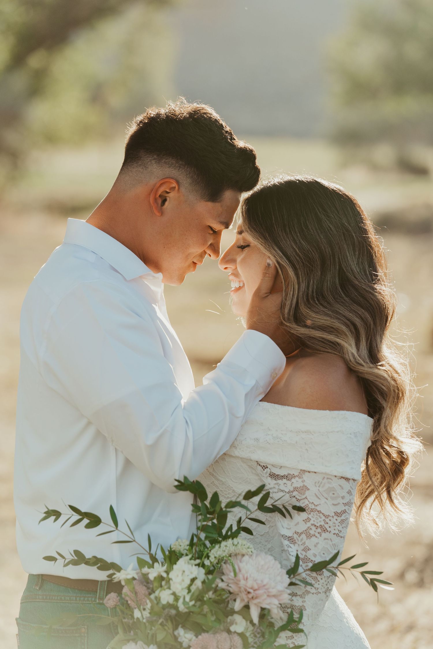 A couple in wedding attire shares a romantic forehead touch outdoors, holding a soft pink floral bouquet in warm golden light.