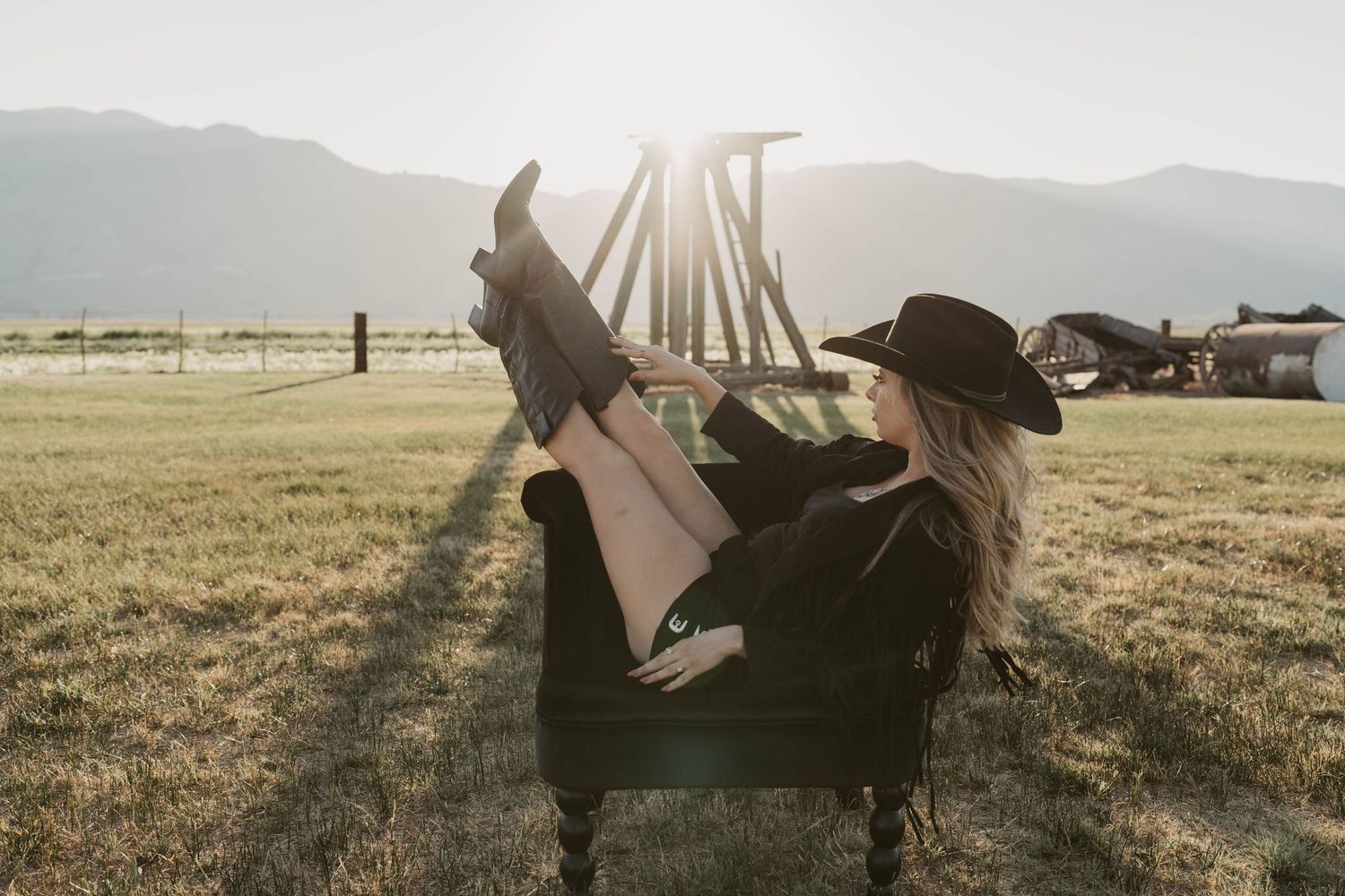 Silhouetted figure relaxes in armchair at sunset in grassy field with mountains in background.