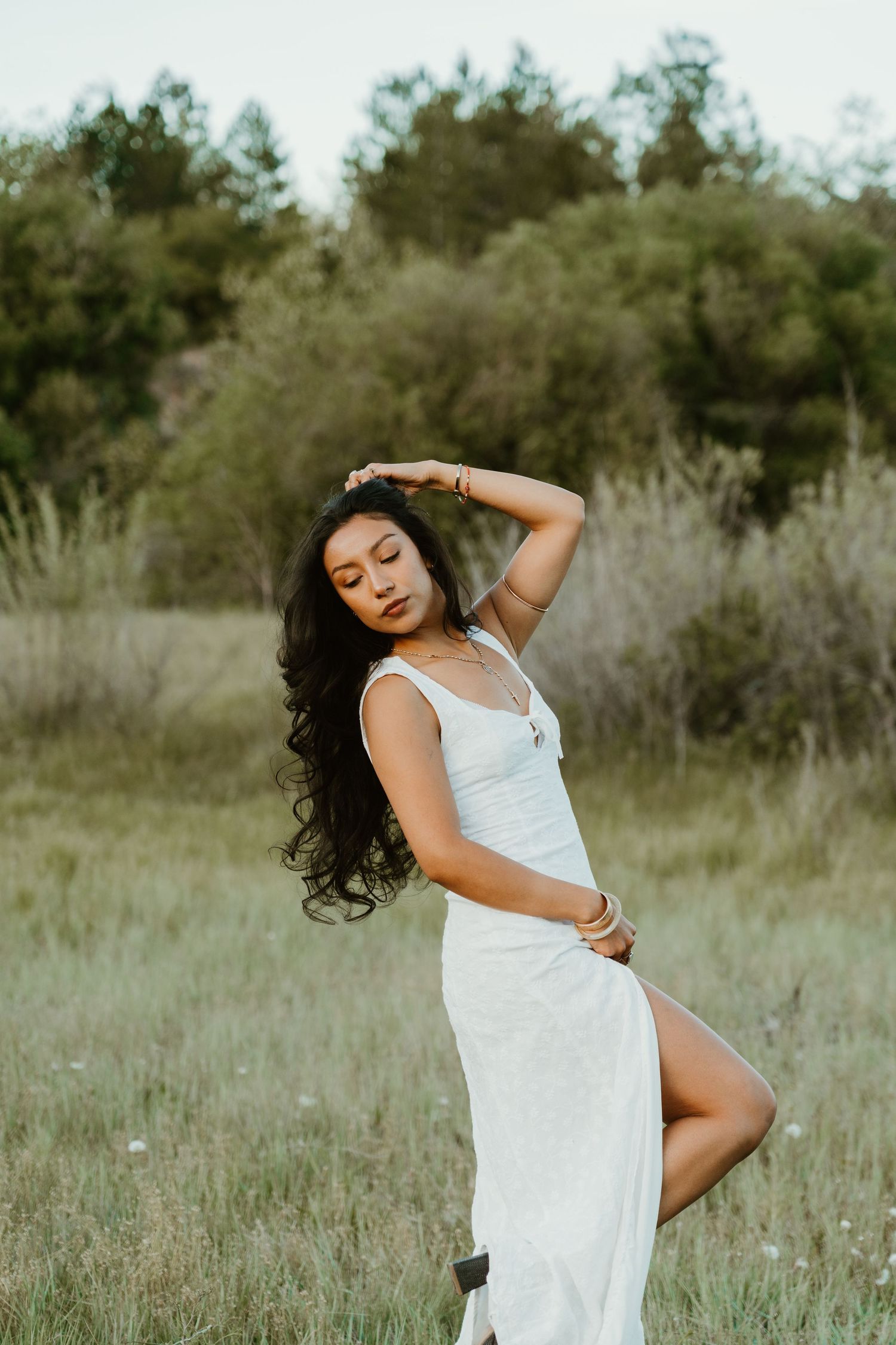 Woman in white slit dress posing gracefully in a green field with hand raised to her hair.
