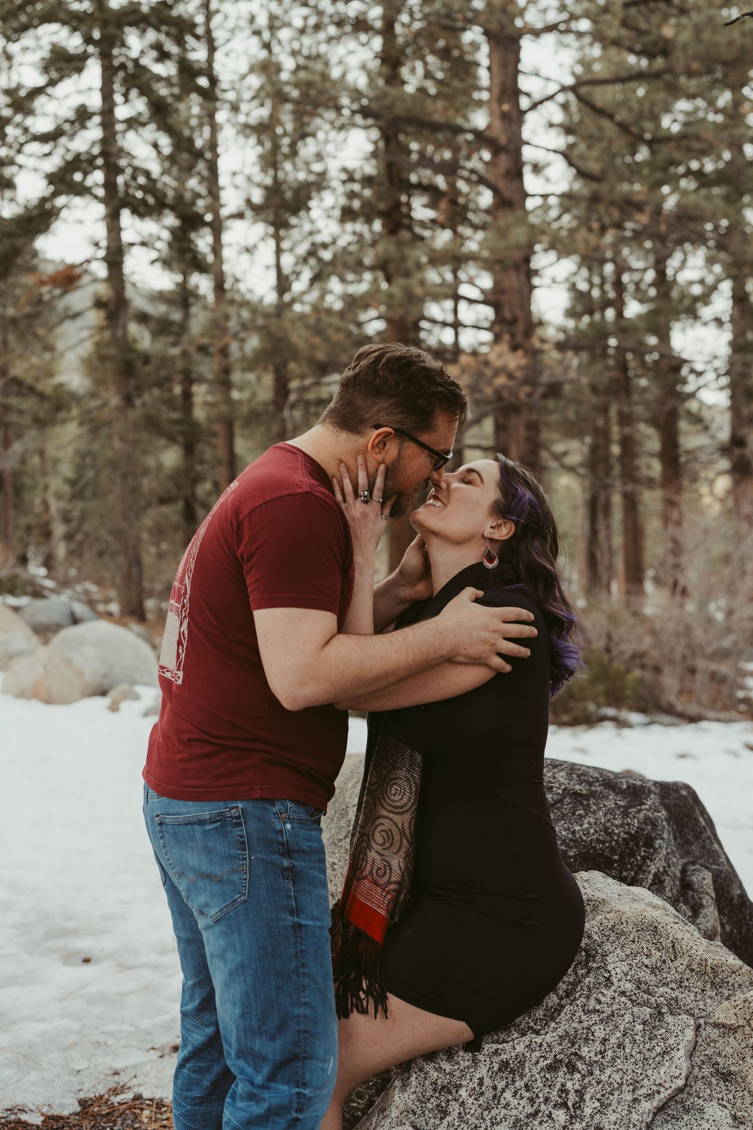 A couple shares a romantic moment on a snow-covered rock surrounded by pine trees in a winter forest setting.