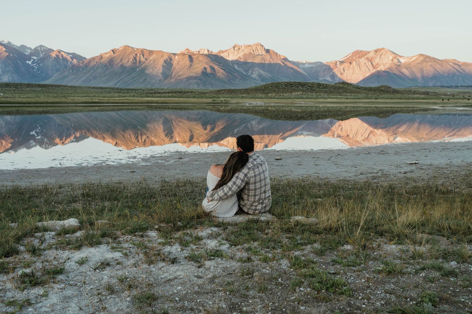 Someone sits by a serene mountain lake at sunset with snow-capped peaks reflecting in the still water.