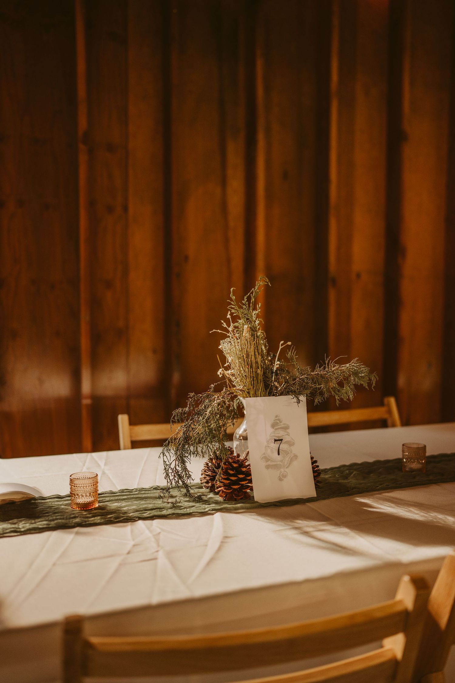 Rustic wedding table setup with pinecone centerpiece, wooden chairs and white tablecloth against warm wooden wall.