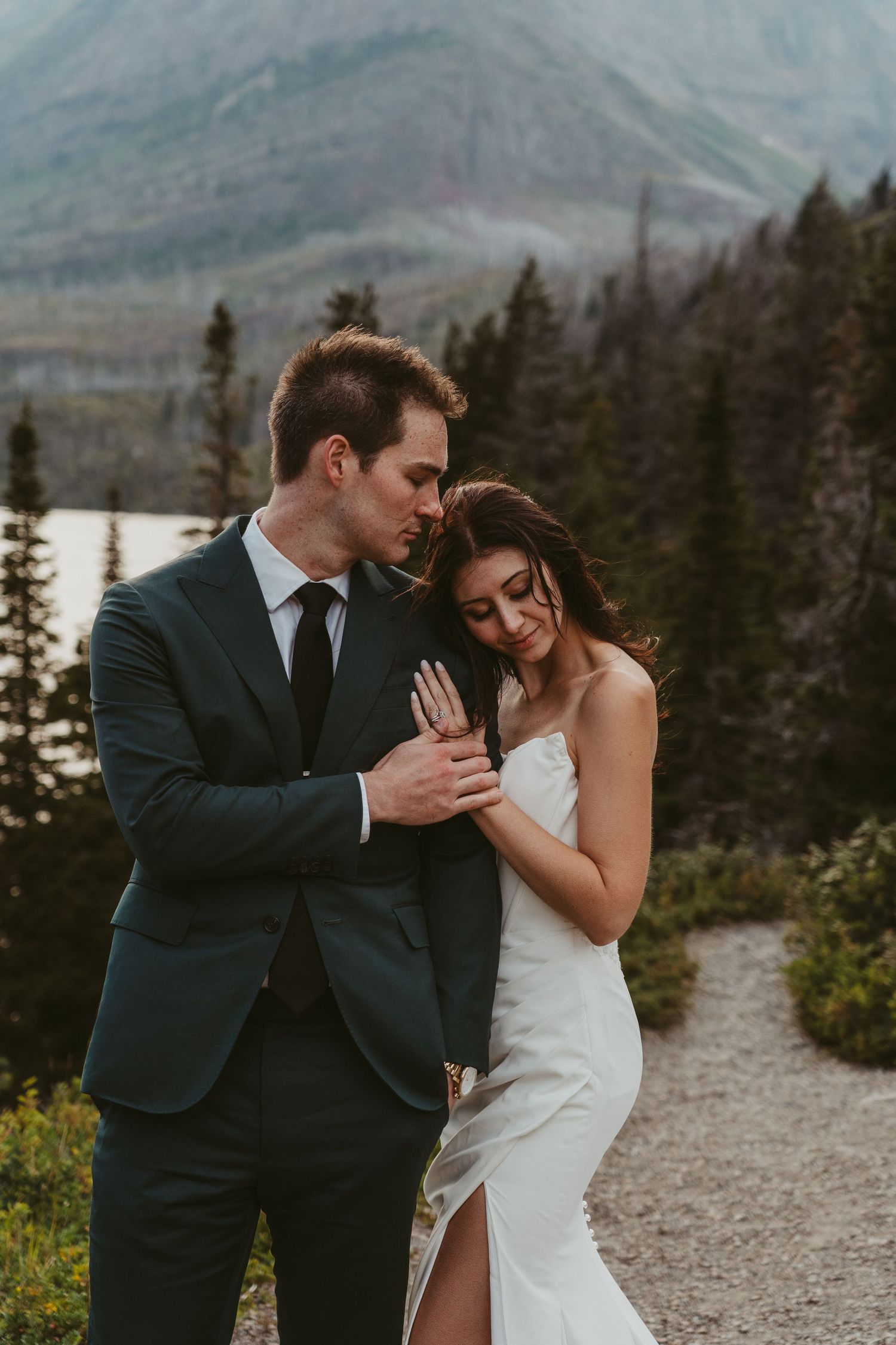 A couple shares an intimate moment on a mountain trail with evergreen trees and misty peaks in the background.