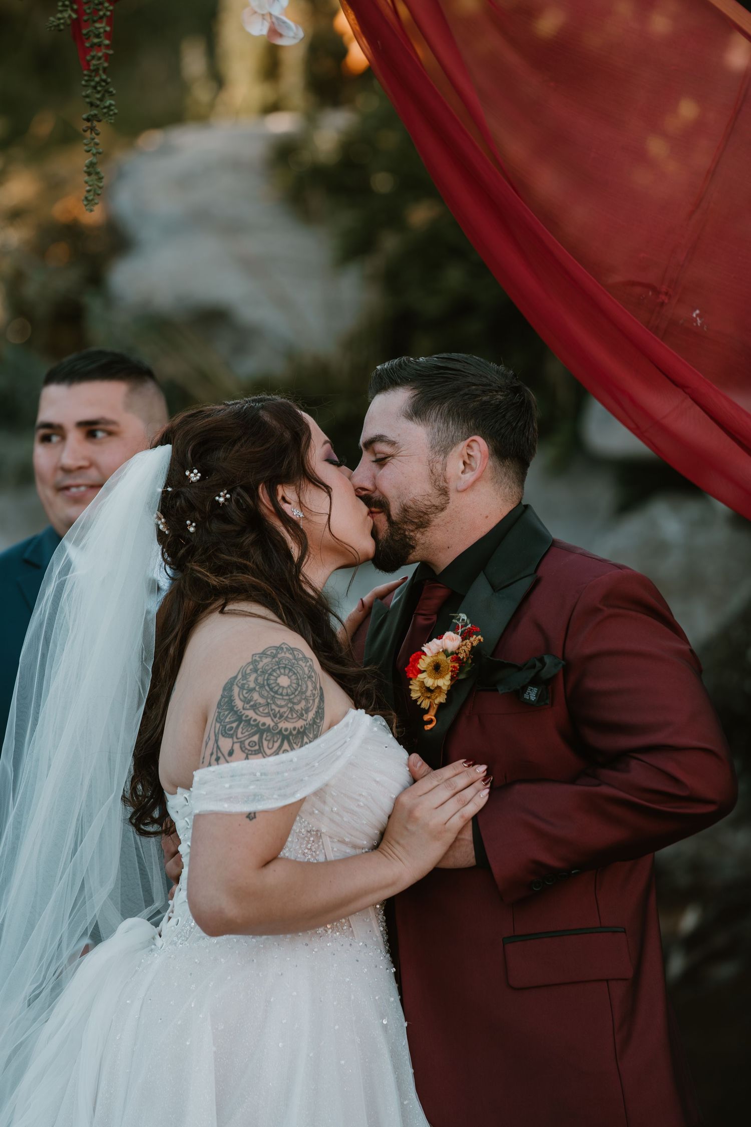 A romantic wedding ceremony kiss between a bride in white lace and groom in burgundy suit under red drapery at sunset.