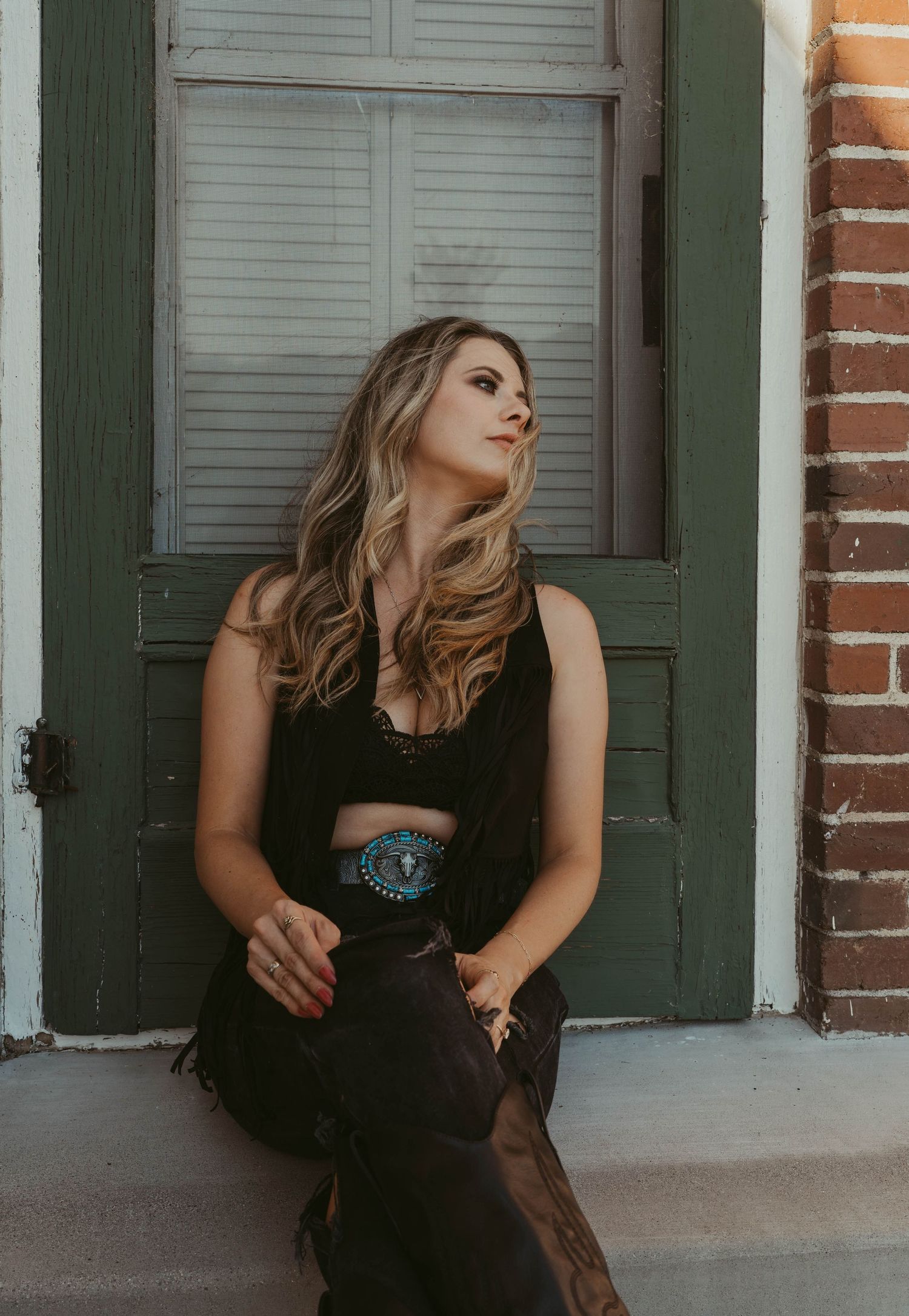 Portrait in black attire against green door and brick wall showcasing western style with vintage belt detail.