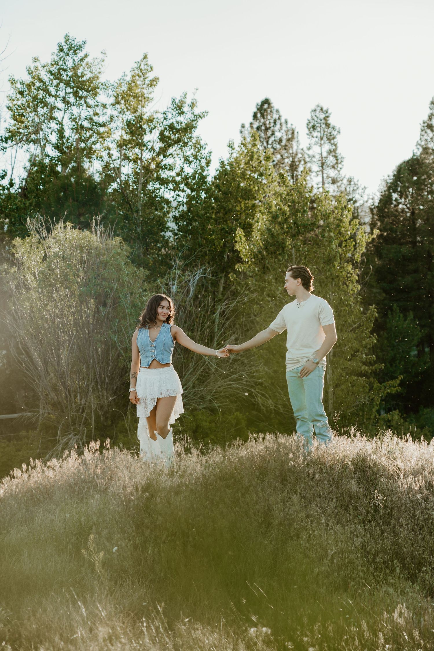 Couple holding hands in a sunlit meadow surrounded by tall grass and green trees during a romantic outdoor photoshoot.