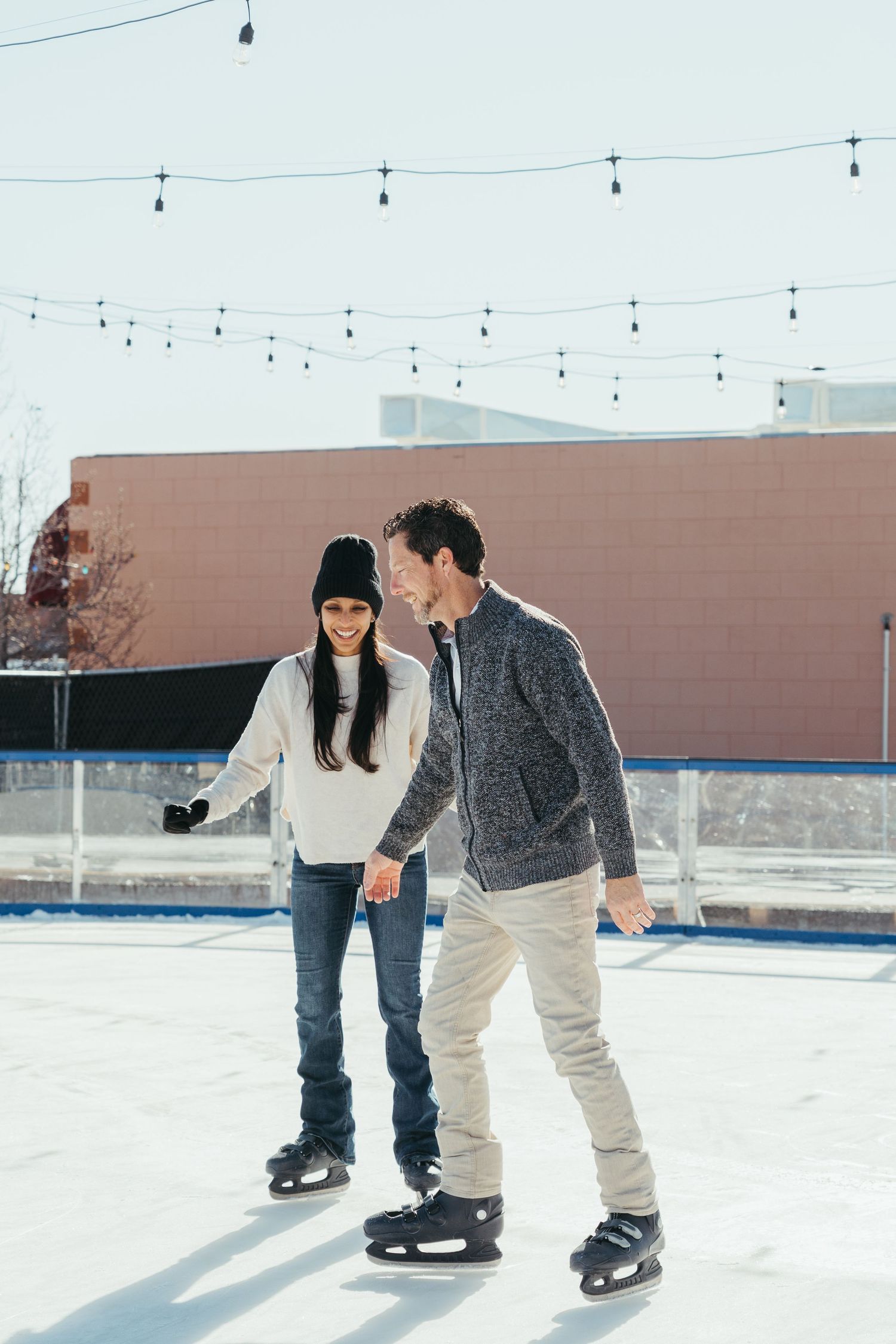 Two people ice skating together on an outdoor rink with string lights overhead and brick buildings in the background.