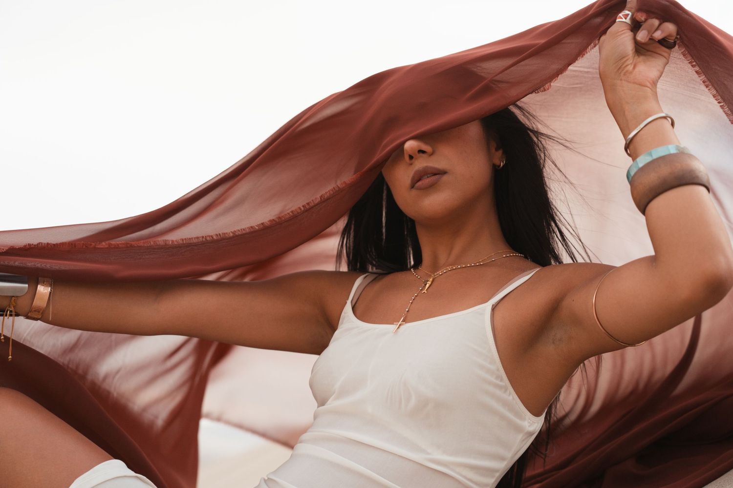Woman in white tank top holding a sheer burgundy fabric over her head outdoors.