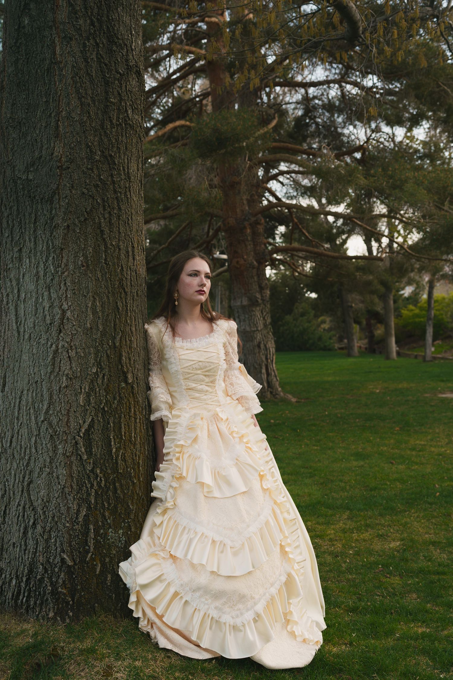 Woman in vintage cream ruffled Victorian gown leaning against large tree trunk in lush green park setting.