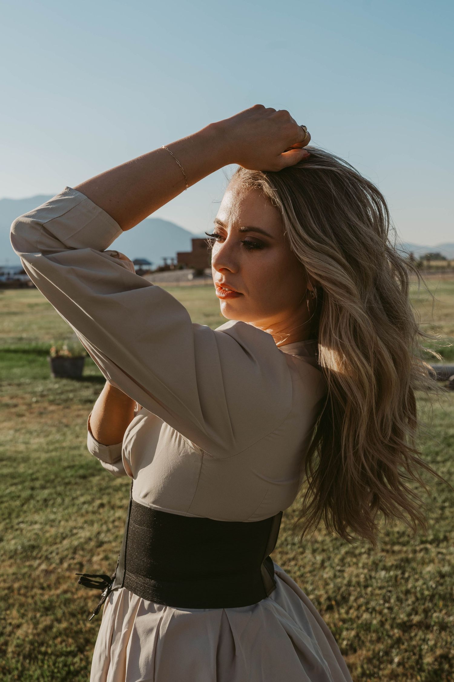 Person with flowing blonde hair poses in tan dress with black corset belt against rural landscape at sunset.