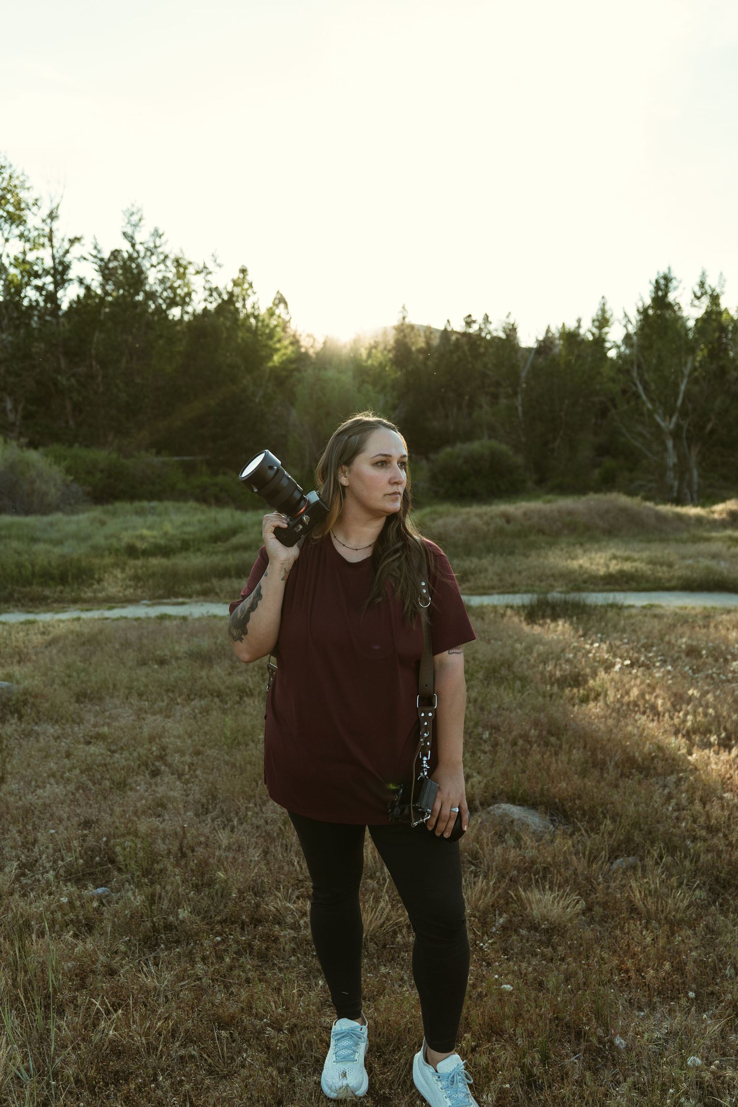 Woman in maroon shirt holds camera on shoulder at golden hour in a grassy field with trees in background.