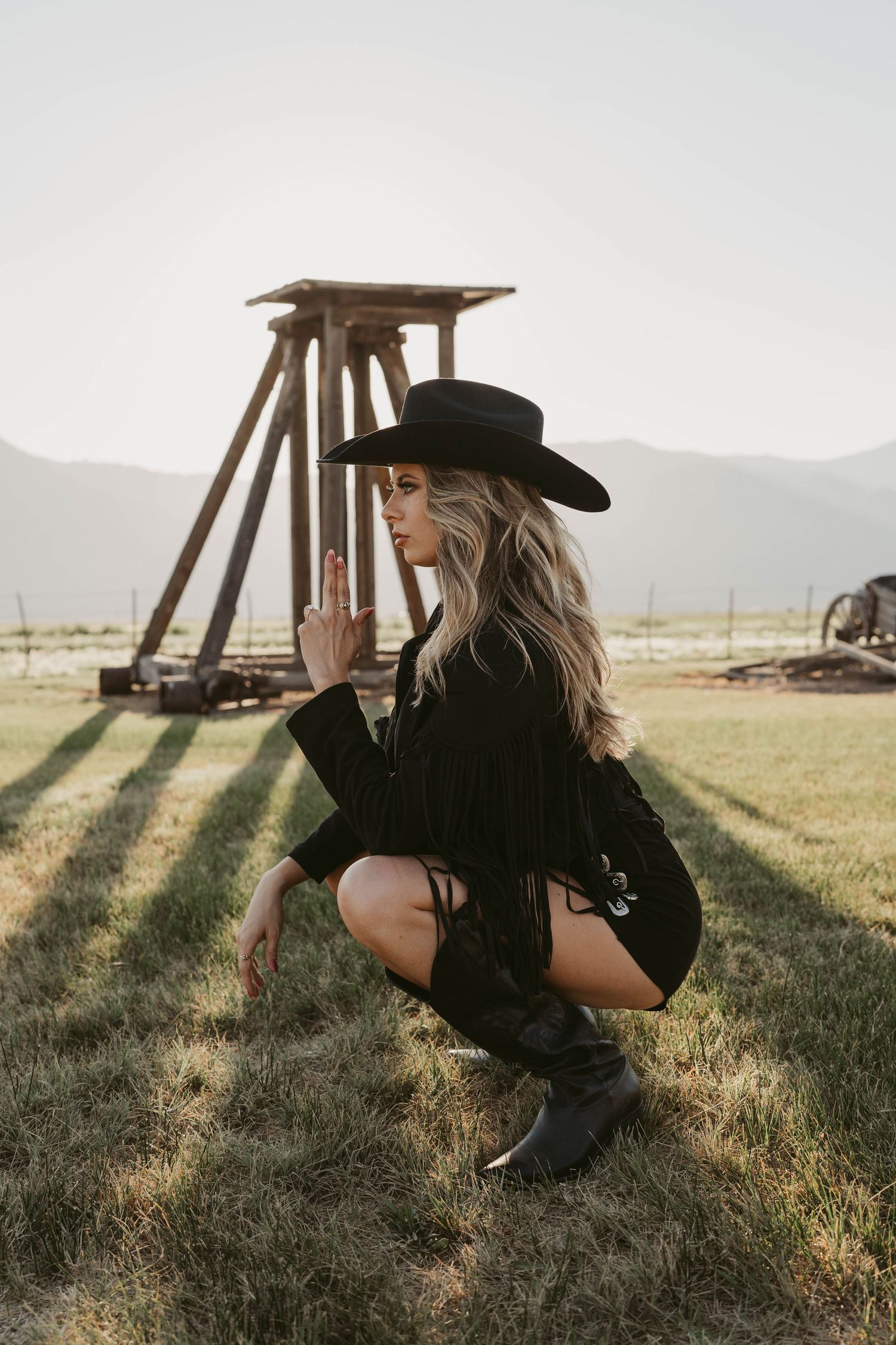 Person in black clothing and wide-brimmed hat crouches in a grassy field at sunset with mountains in the distance.